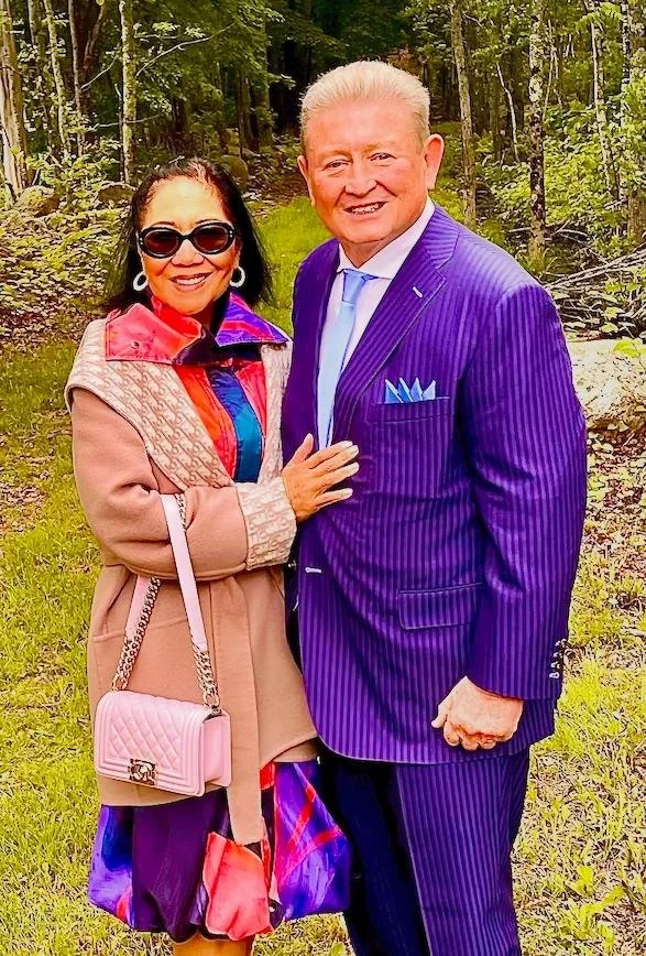 A smiling woman wearing sunglasses and a beige coat standing next to a smiling man in a purple pinstripe suit in a wooded outdoor setting — Throwback Dating singles events Chicago over 50.