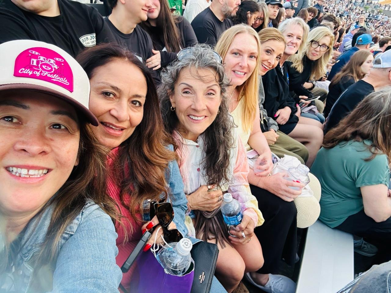 Group of smiling people sitting in stadium seats at an outdoor event, some holding water bottles, with more attendees visible in the background.