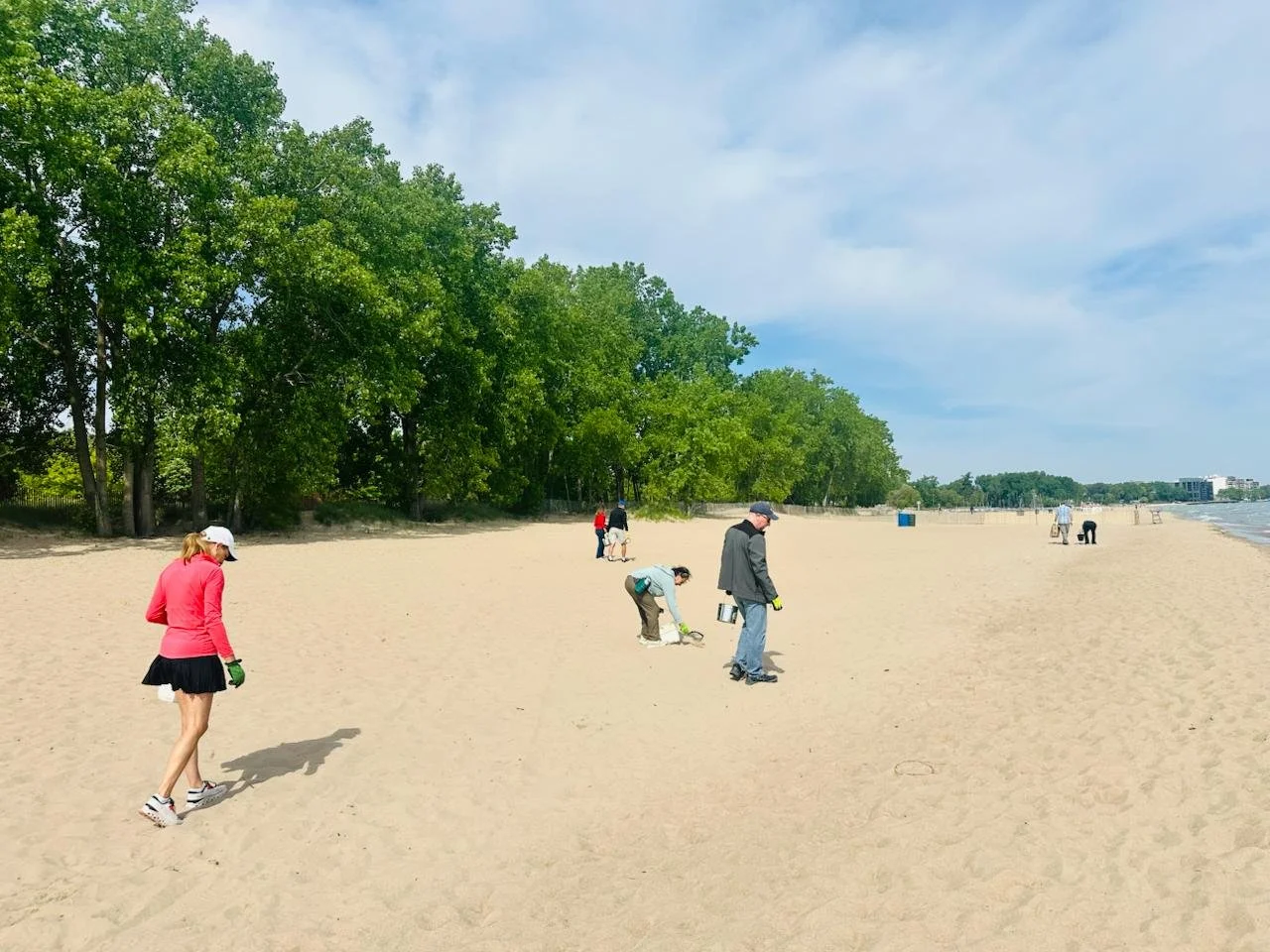 People cleaning up trash on a sandy beach with trees and buildings in the background on a partly cloudy day — Throwback Dating singles events Chicago over 50.