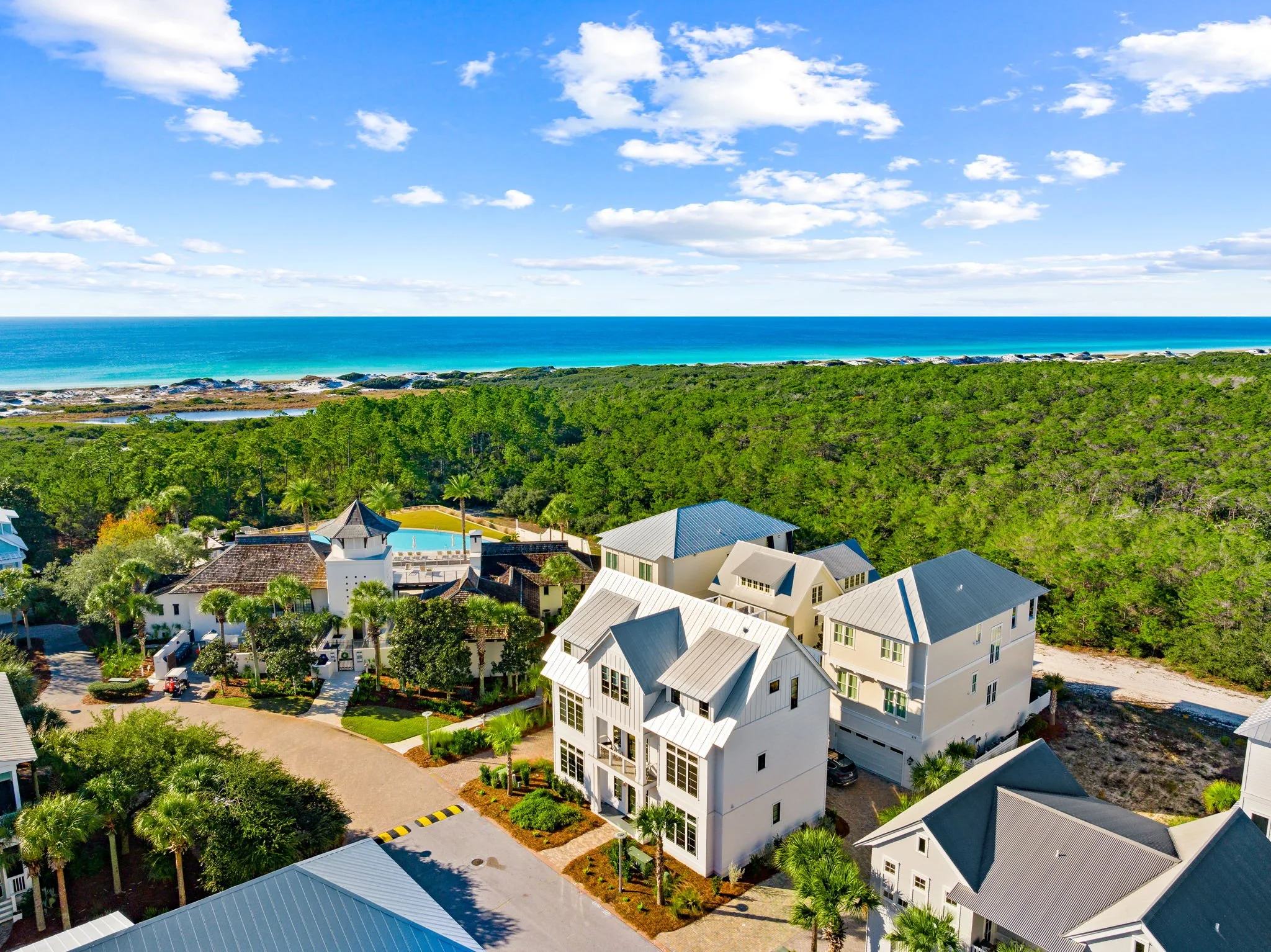 Aerial view of a coastal area with white houses, lush green trees, a swimming pool, and the beach and ocean in the background with blue sky and scattered clouds.