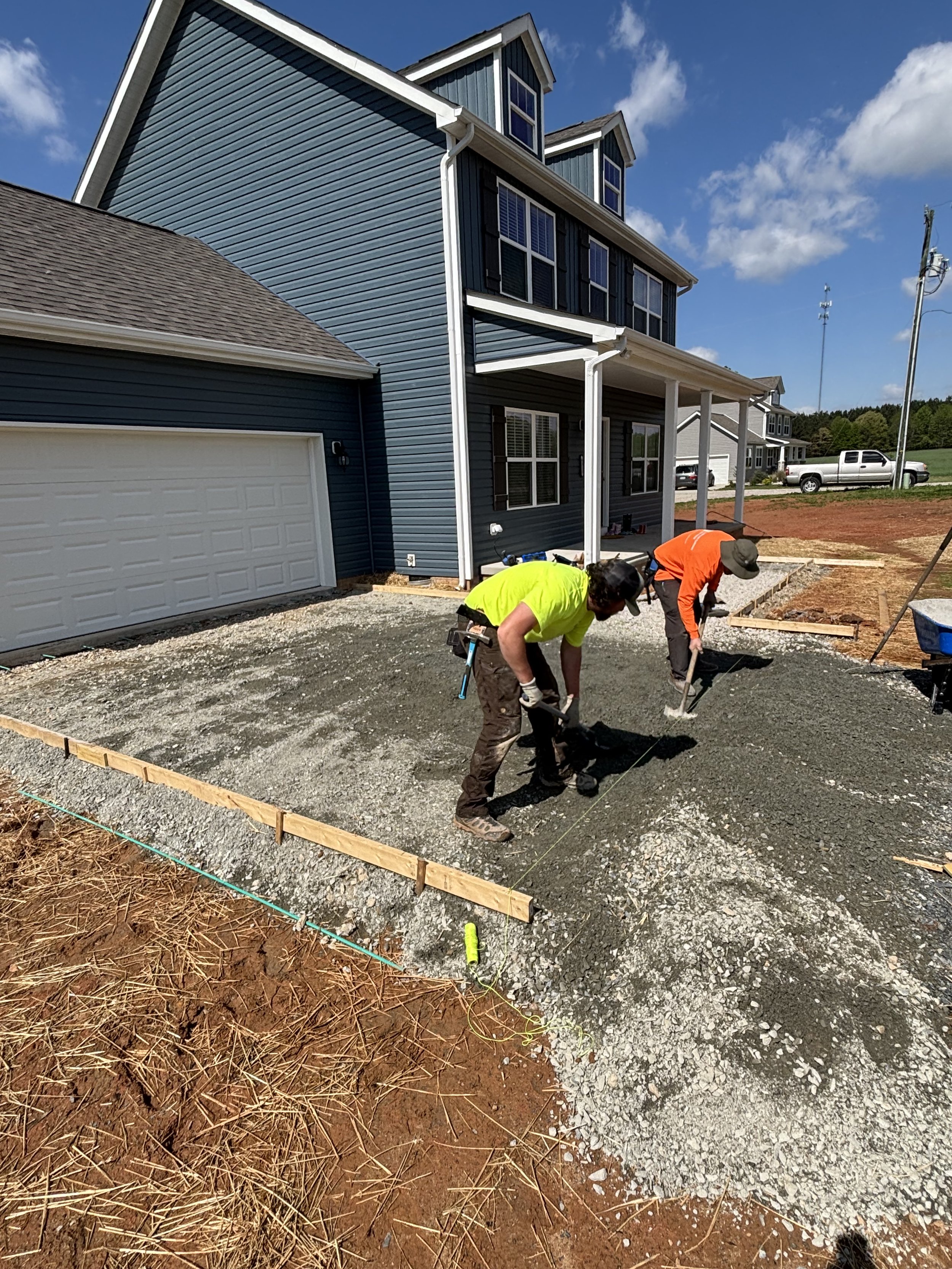 Two construction workers preparing ground for concrete driveway in front of two-story blue house.