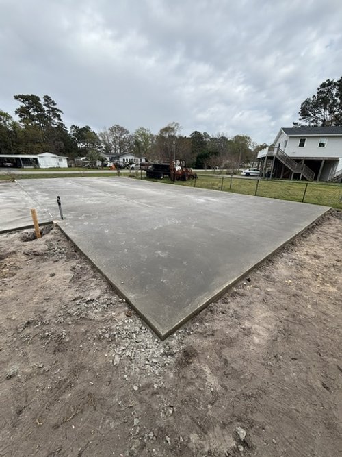 Freshly poured concrete foundation slab in a residential area with houses and grassy lawns in the background.