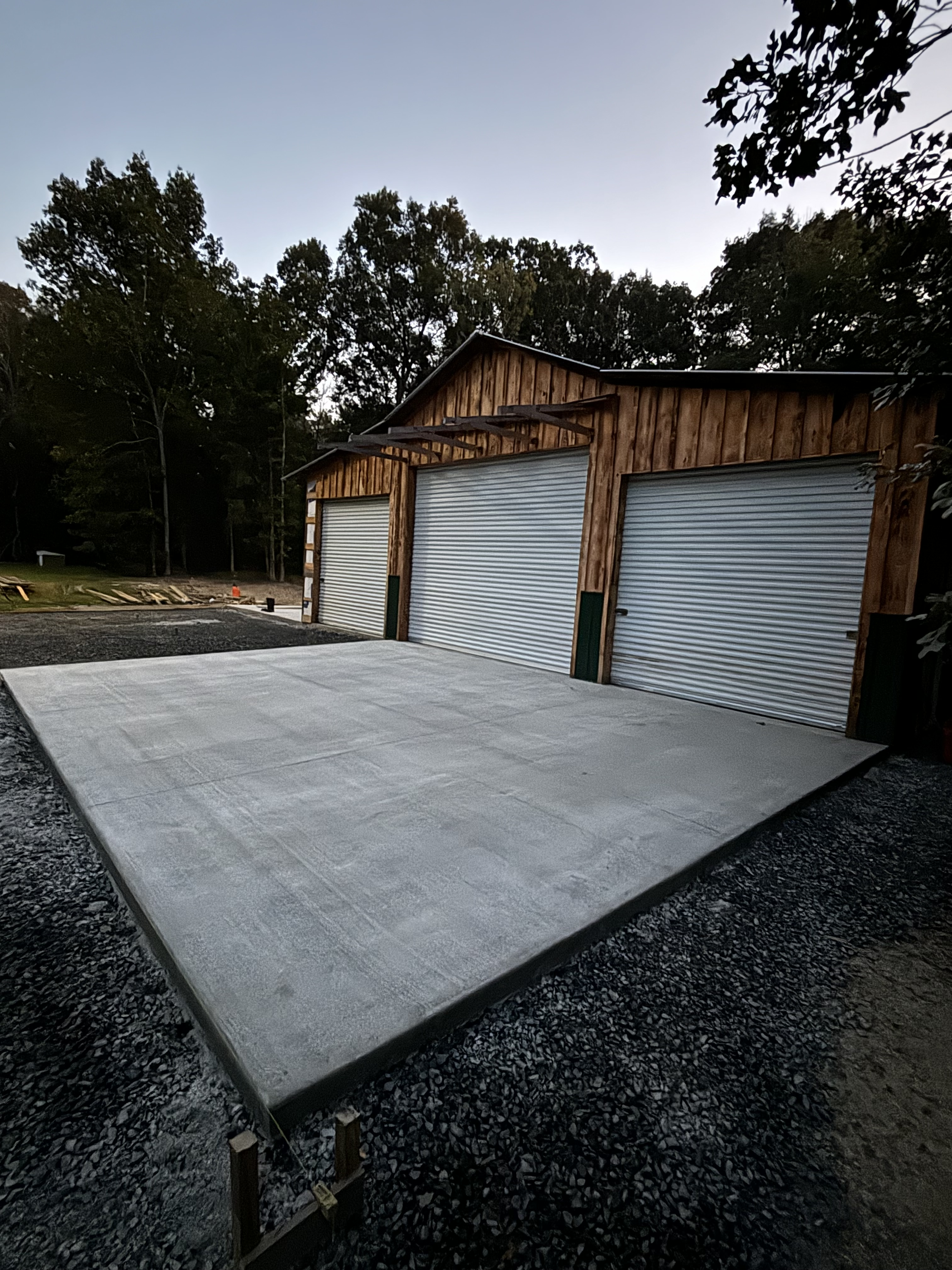 A large wooden garage with three metal roll-up doors and a freshly poured concrete slab in front, surrounded by trees and gravel.