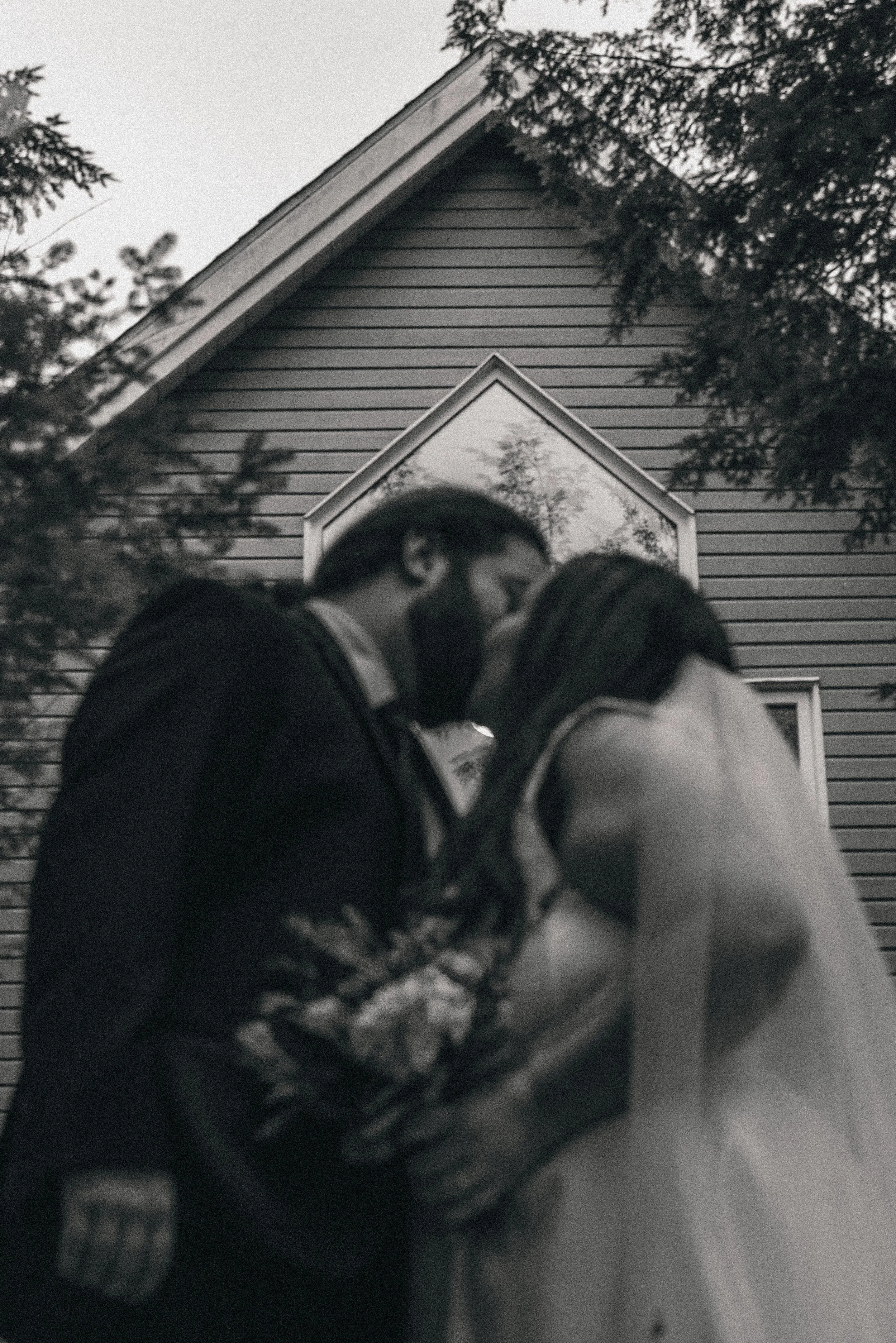A couple is kissing outdoors in front of a house with siding and windows, surrounded by trees.