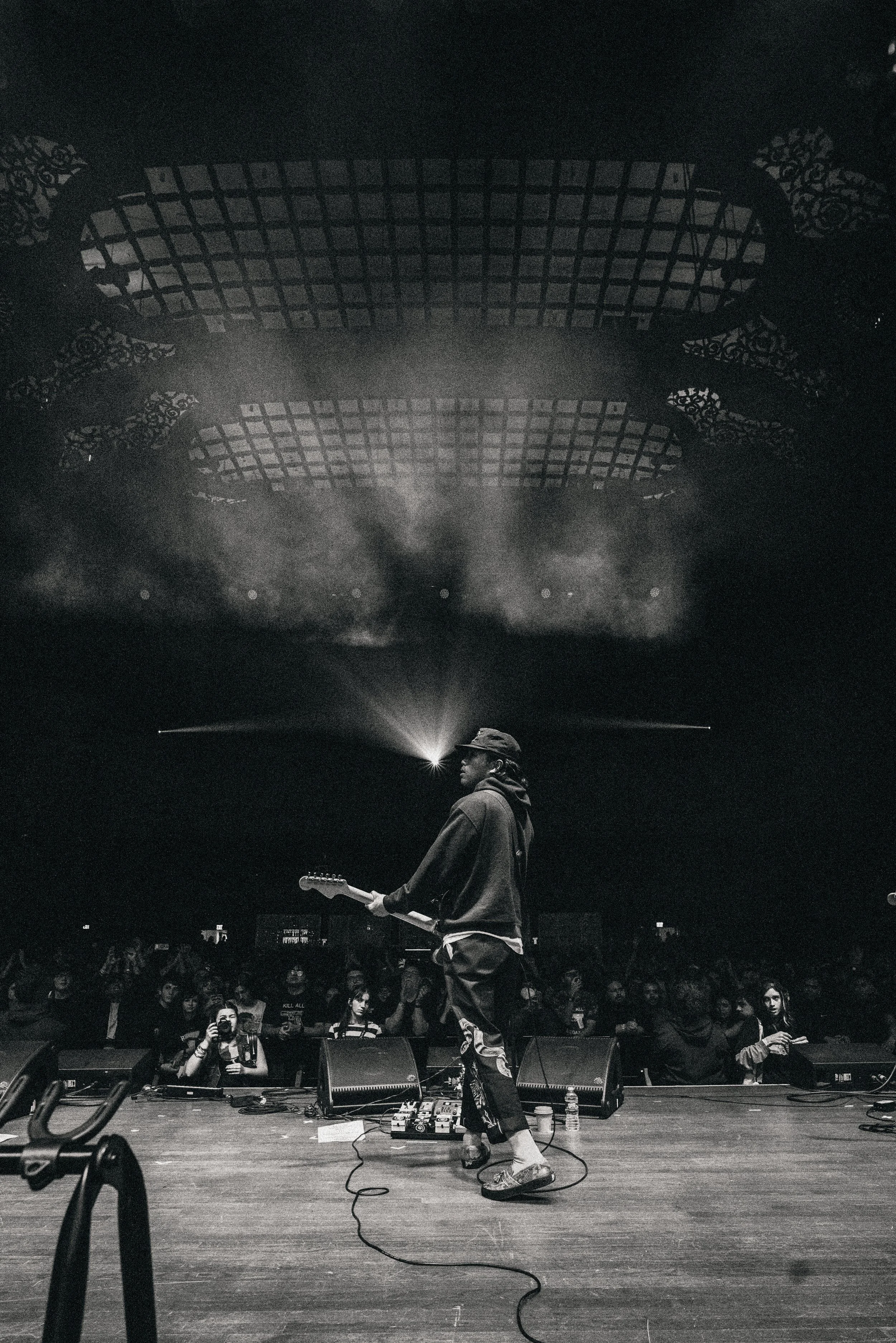 A musician plays guitar on stage in front of an audience, with dramatic lighting and a decorative ceiling above.