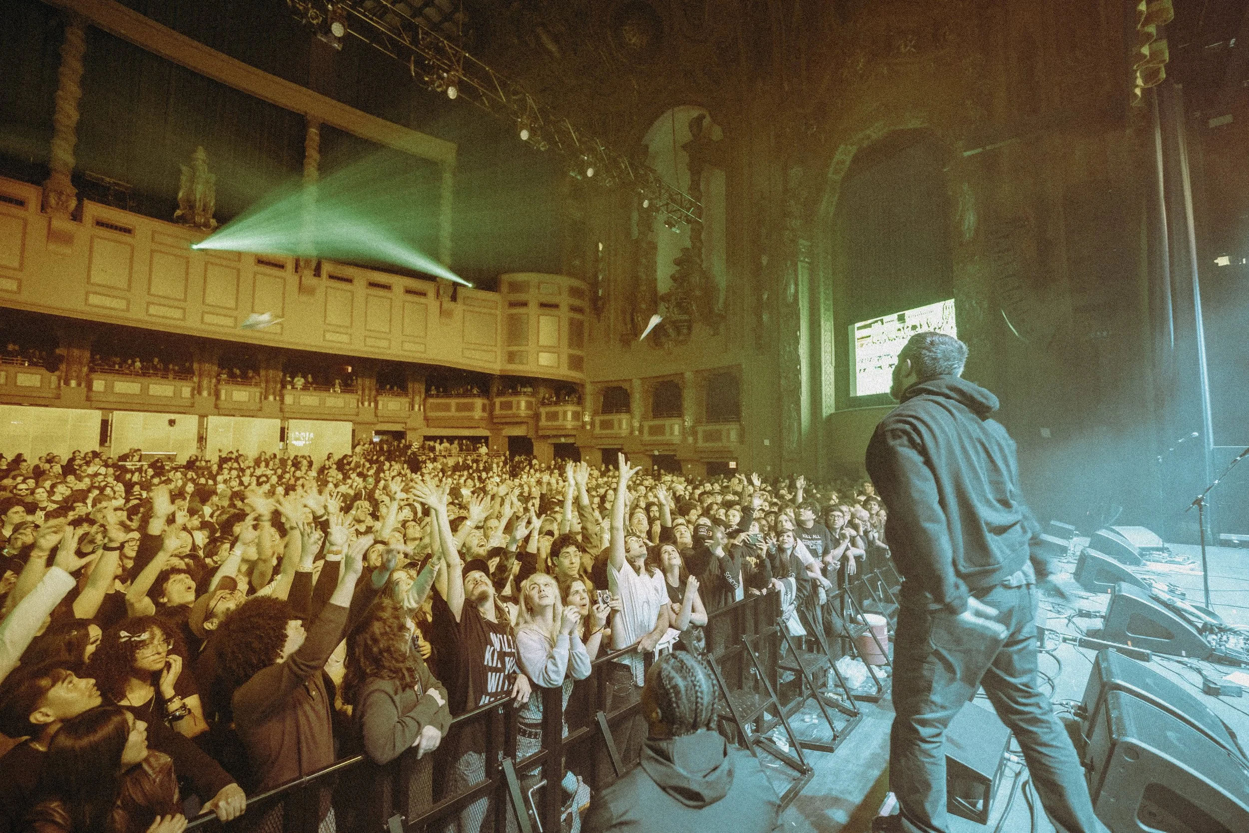 A performer on stage with his back to the camera in front of a large crowd at a concert venue.