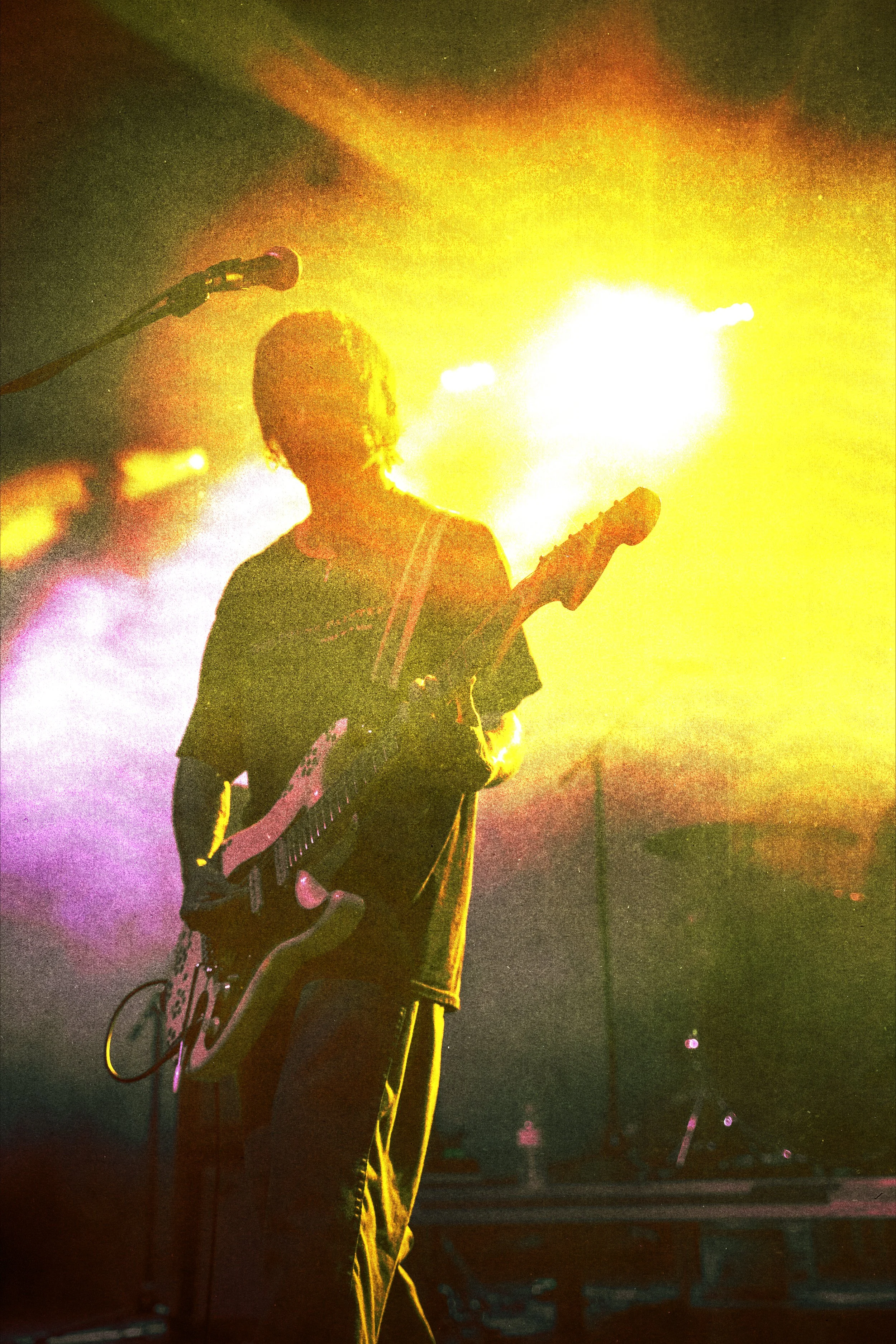 A person playing an electric guitar on stage, silhouetted by bright yellow and orange stage lights, with a microphone on a stand nearby.