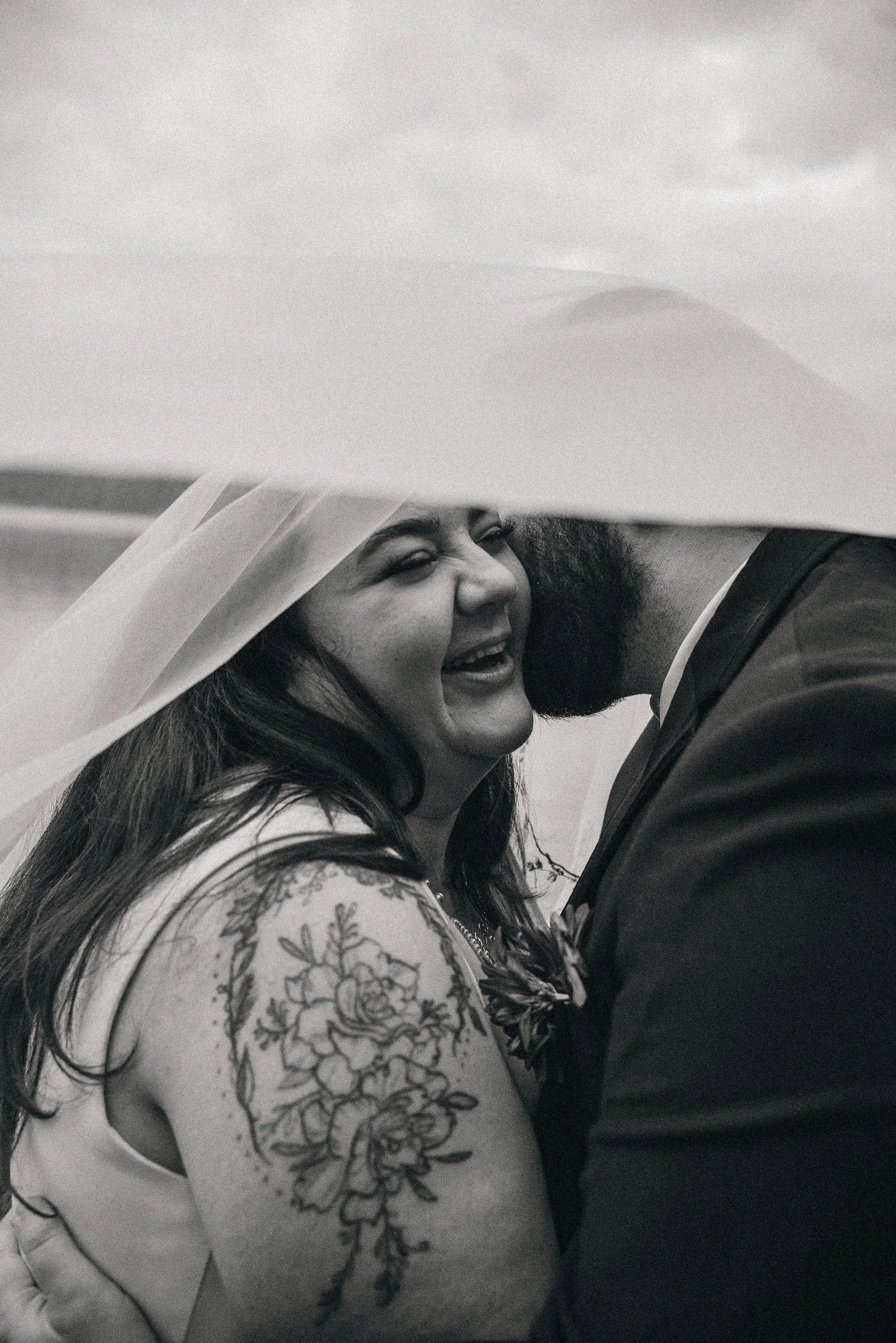 Black and white photograph of a couple sharing an intimate moment under a veil during their wedding, with the bride smiling and displaying a floral tattoo on her arm.