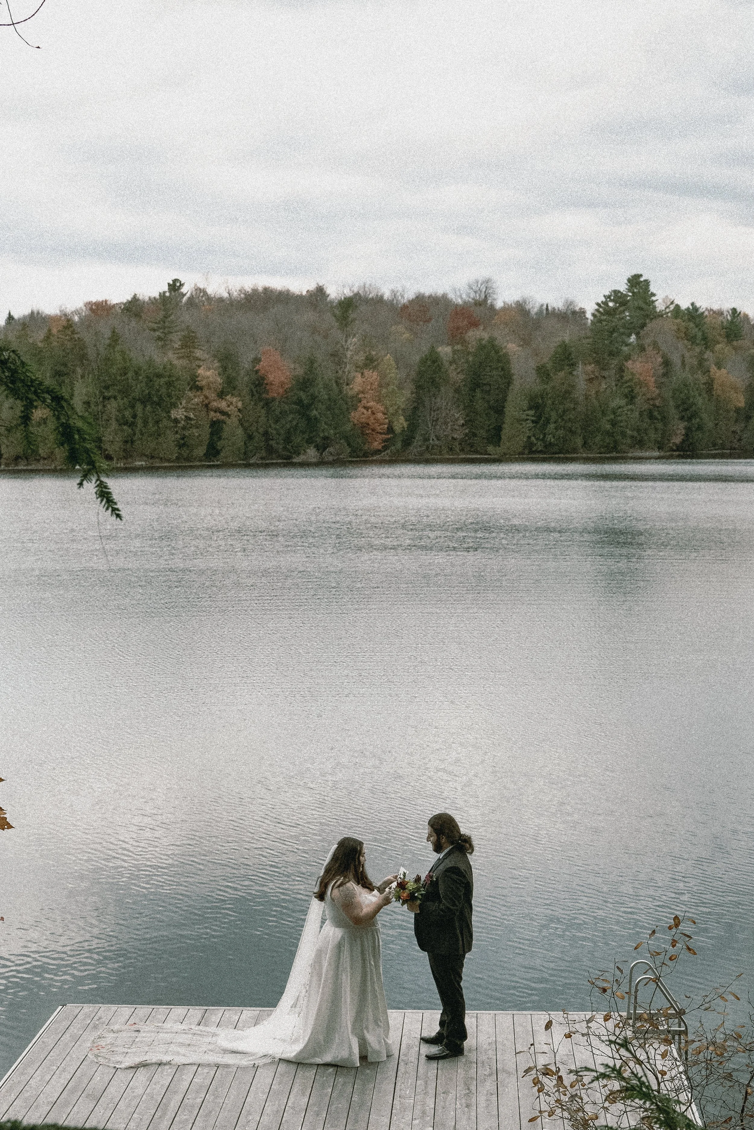 A couple exchanging vows on a wooden dock by a lake with a forested shoreline in the background, overcast sky.