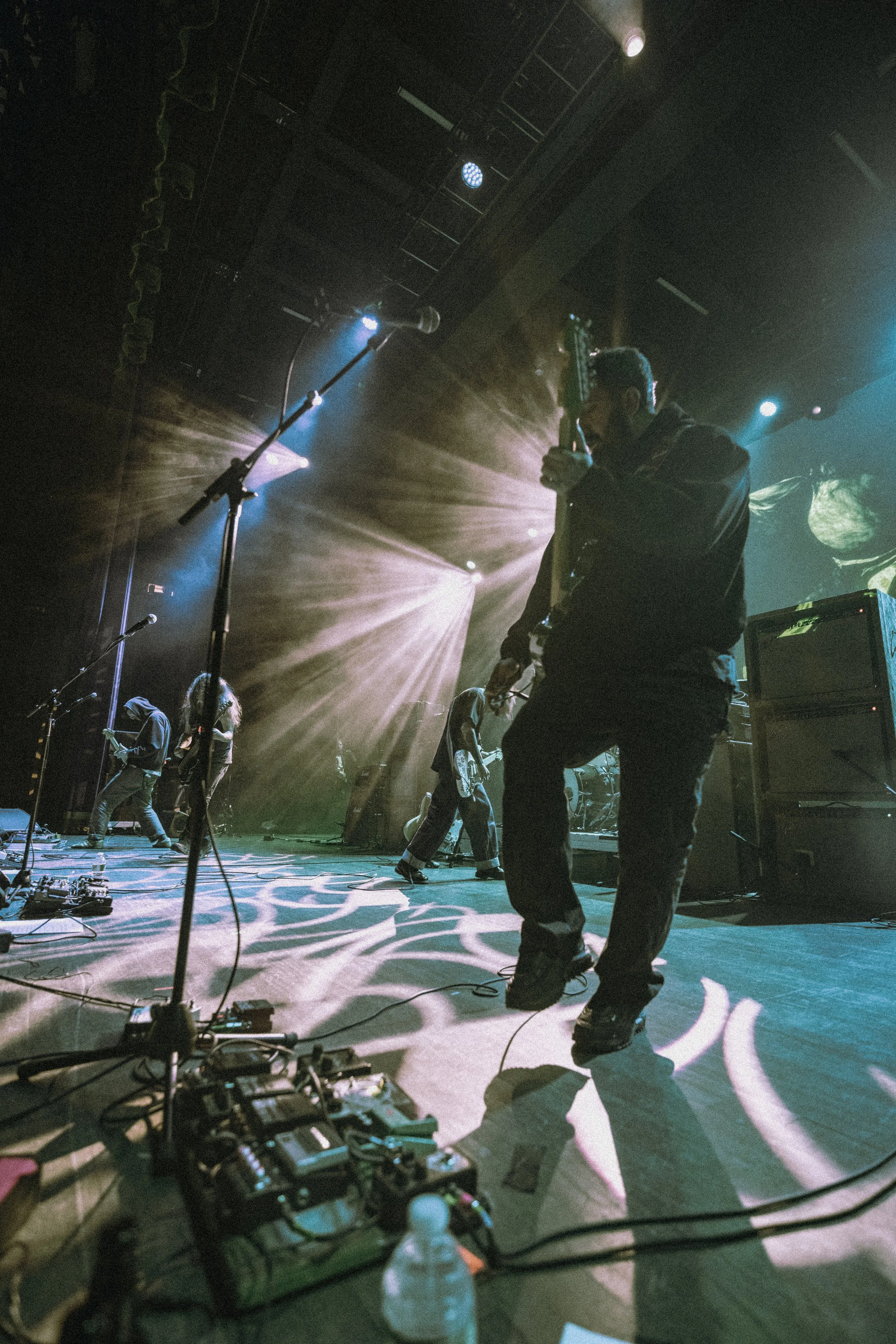 Musicians performing on stage with a guitarist in the foreground, stage lighting, microphone, and music equipment visible.