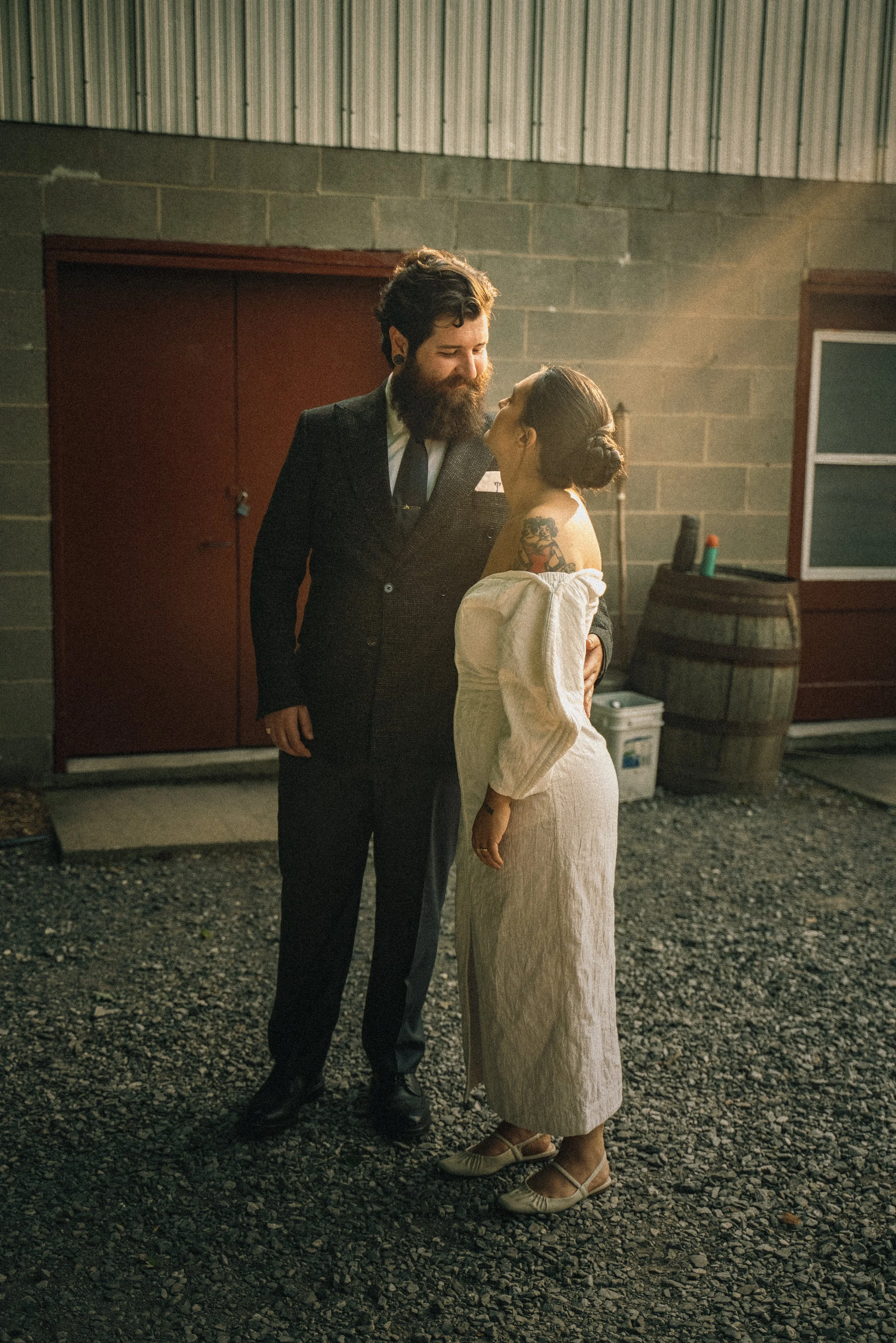A man and woman in vintage clothing sharing a tender moment in an industrial setting with gravel ground and a barrel in the background.