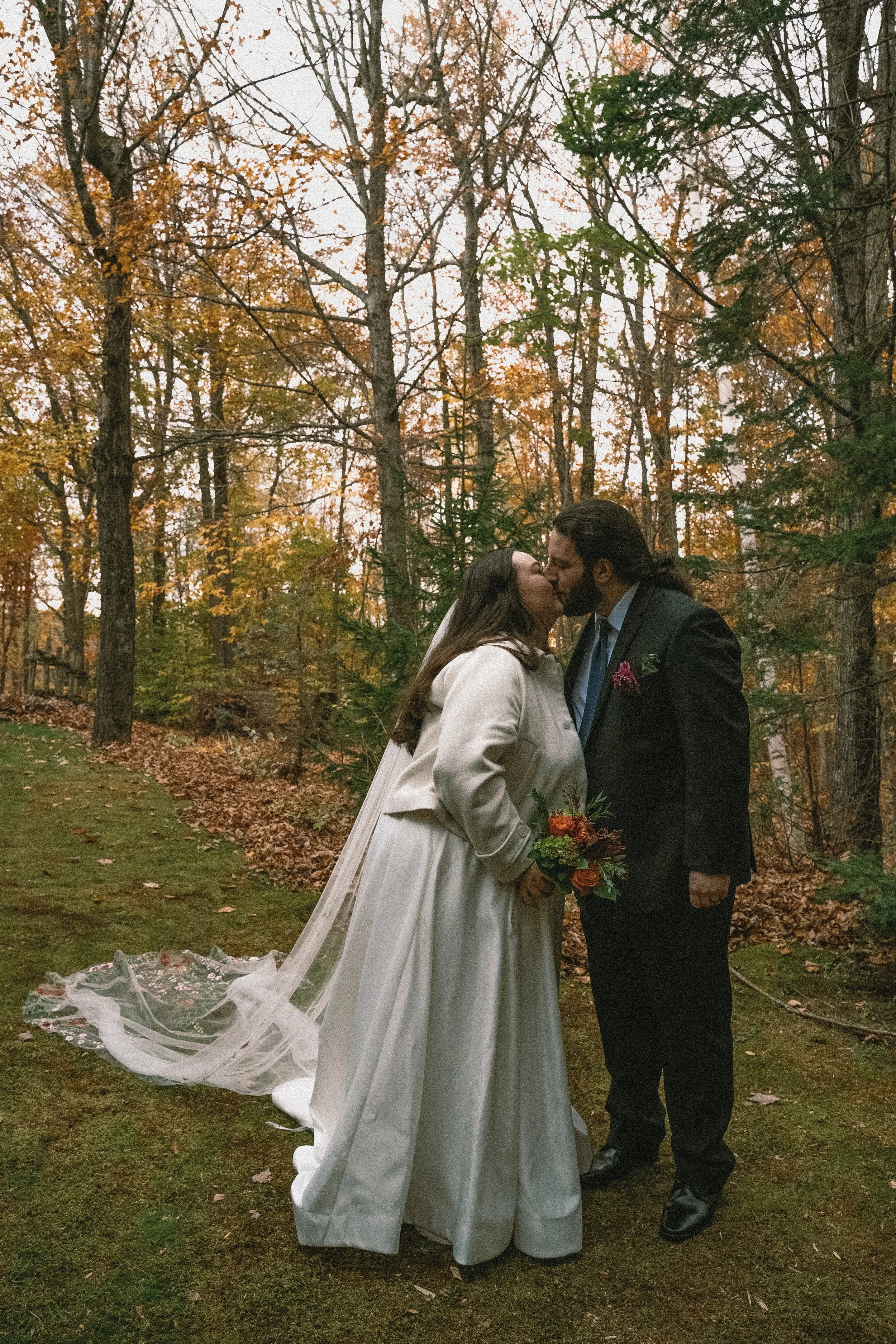 A couple sharing a kiss outdoors in a wooded area on a fall day, with trees and fallen leaves in the background. The woman wears a white wedding dress, and the man wears a dark suit with a light blue shirt and dark tie. The woman holds a bouquet of f