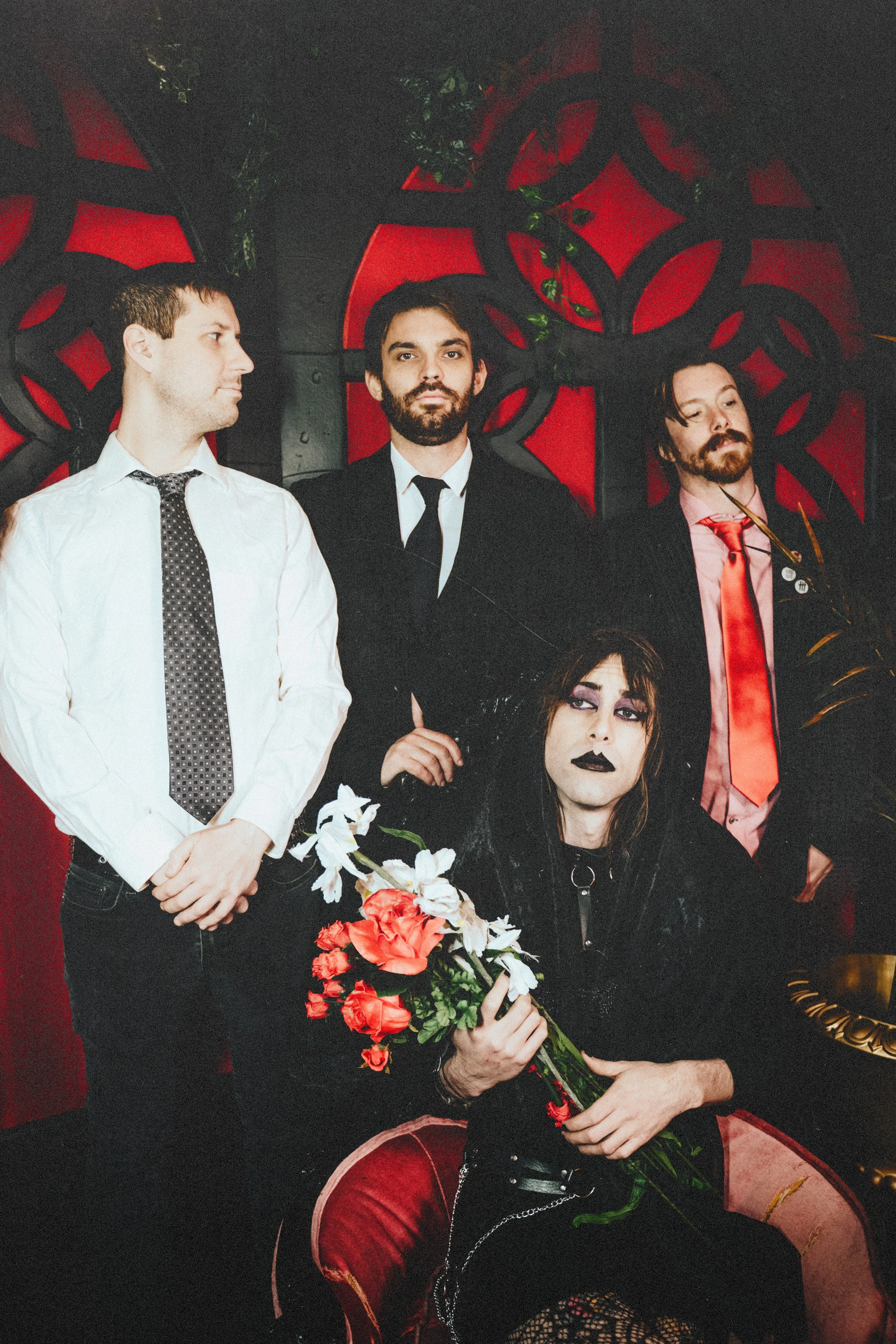 A group of four people in dark and formal clothing pose against a red and black decorative background. The person sitting in front is a woman holding a bouquet of flowers, with dark makeup and long dark hair. The three men stand behind her, with two 