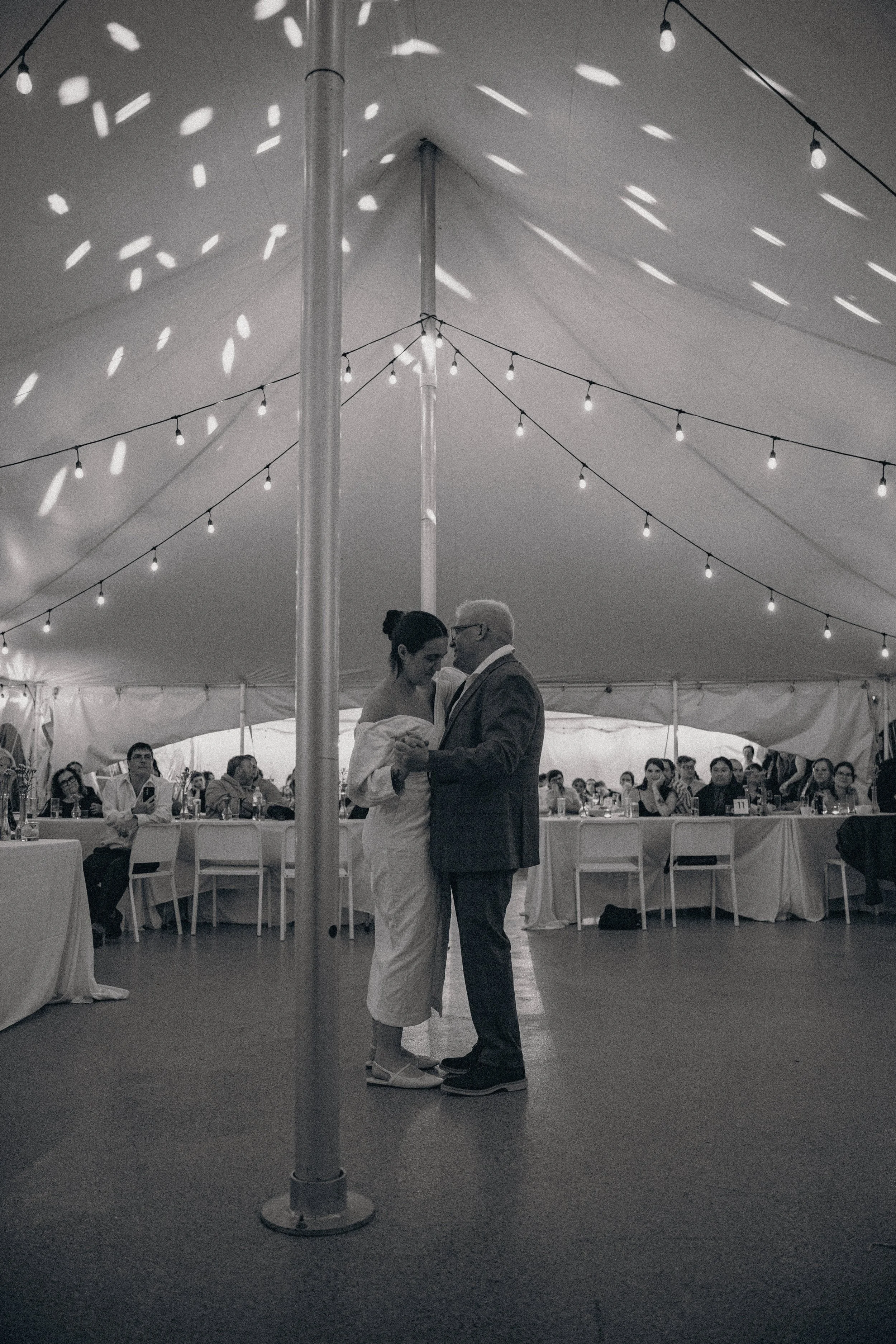 Black and white photo of a bride and groom dancing under a tent with string lights, surrounded by seated guests at tables.