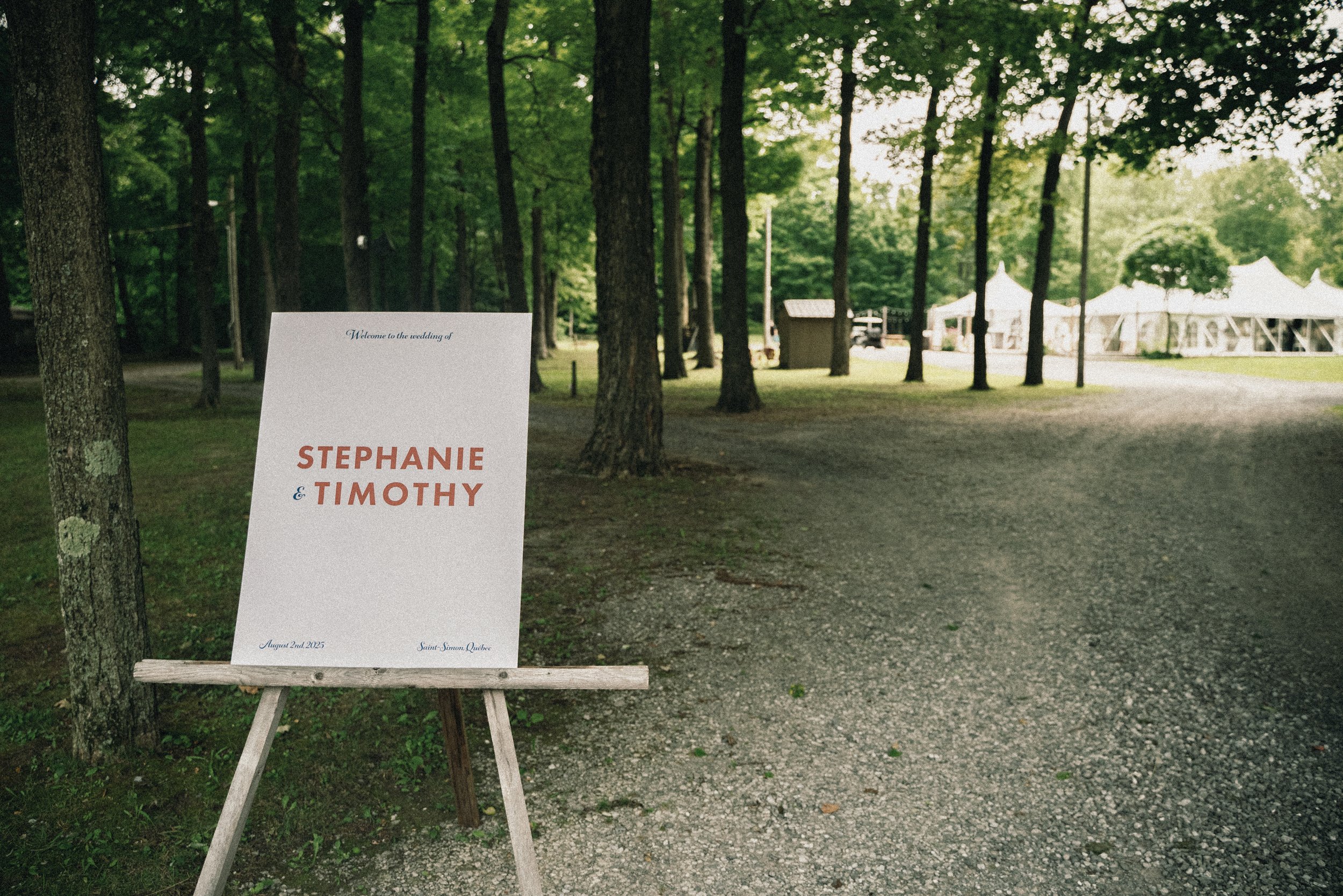 Wedding sign in a wooded outdoor setting, reading 'Welcome to the wedding of Stephanie & Timothy, August 2nd, 2025, Saint-Simon, Quebec', with tents visible in the background.