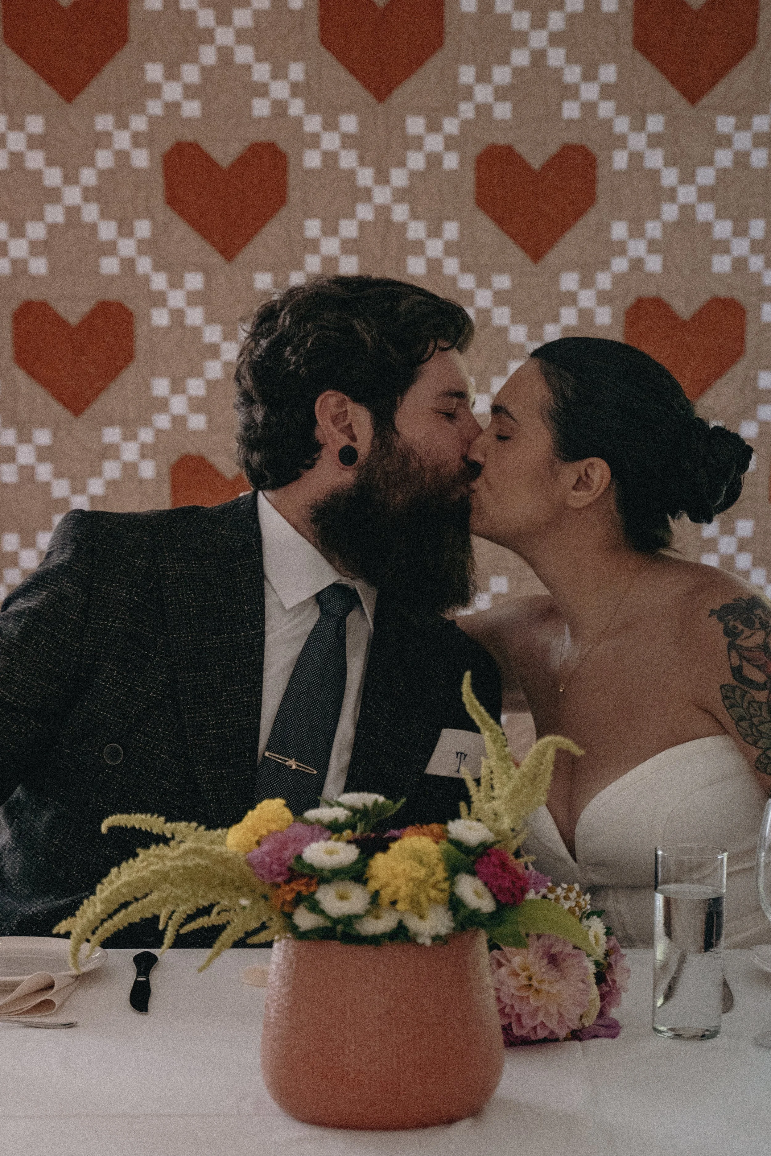 A couple sharing a kiss at a wedding reception, with a background of heart-patterned wallpaper and a flower centerpiece on the table in front of them.