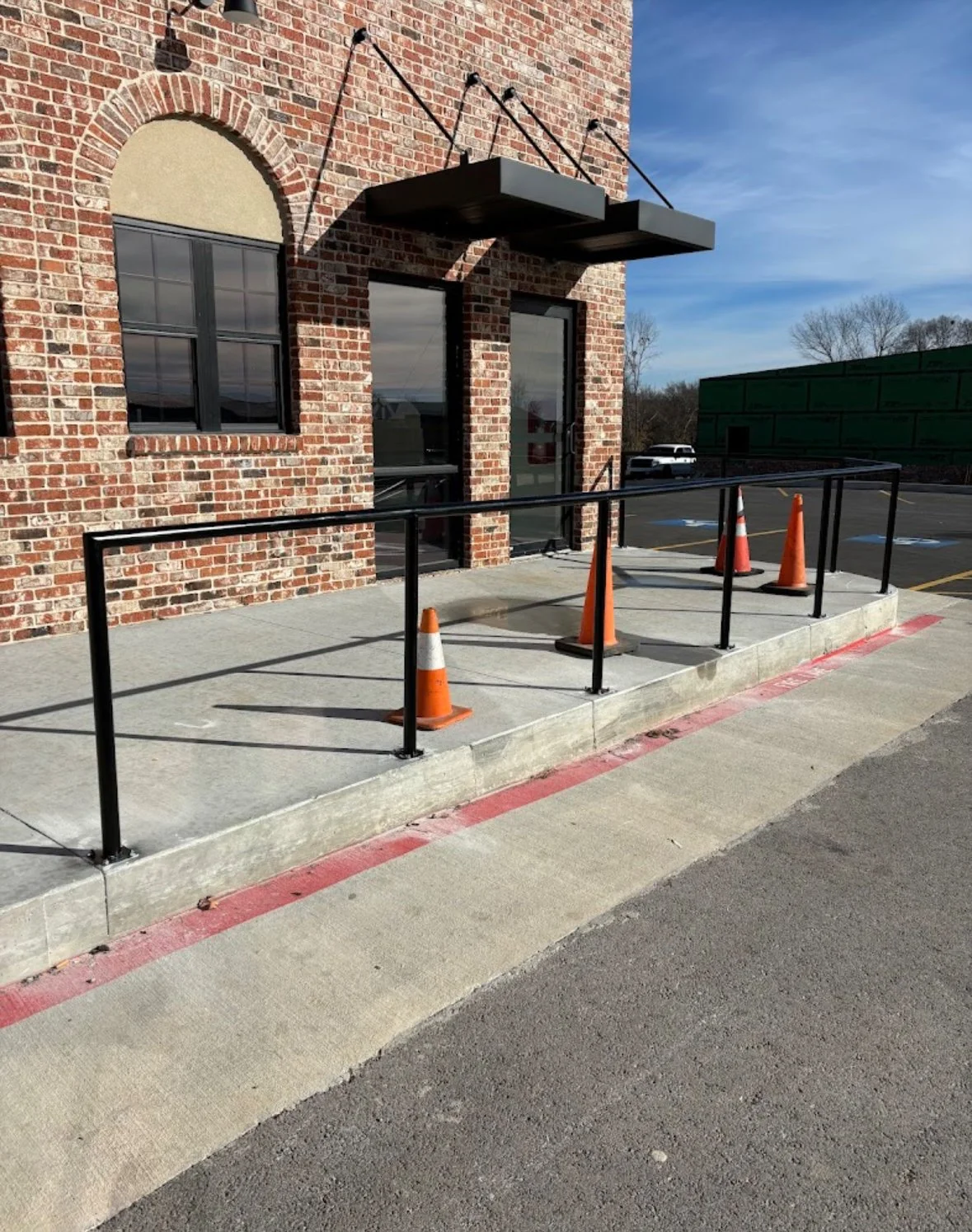 Exterior of a brick building with a glass door, two windows, black metal handrails by A1 Metal Fabrication, and orange traffic cones on a concrete sidewalk. Clear sky and parked cars are visible in the background.