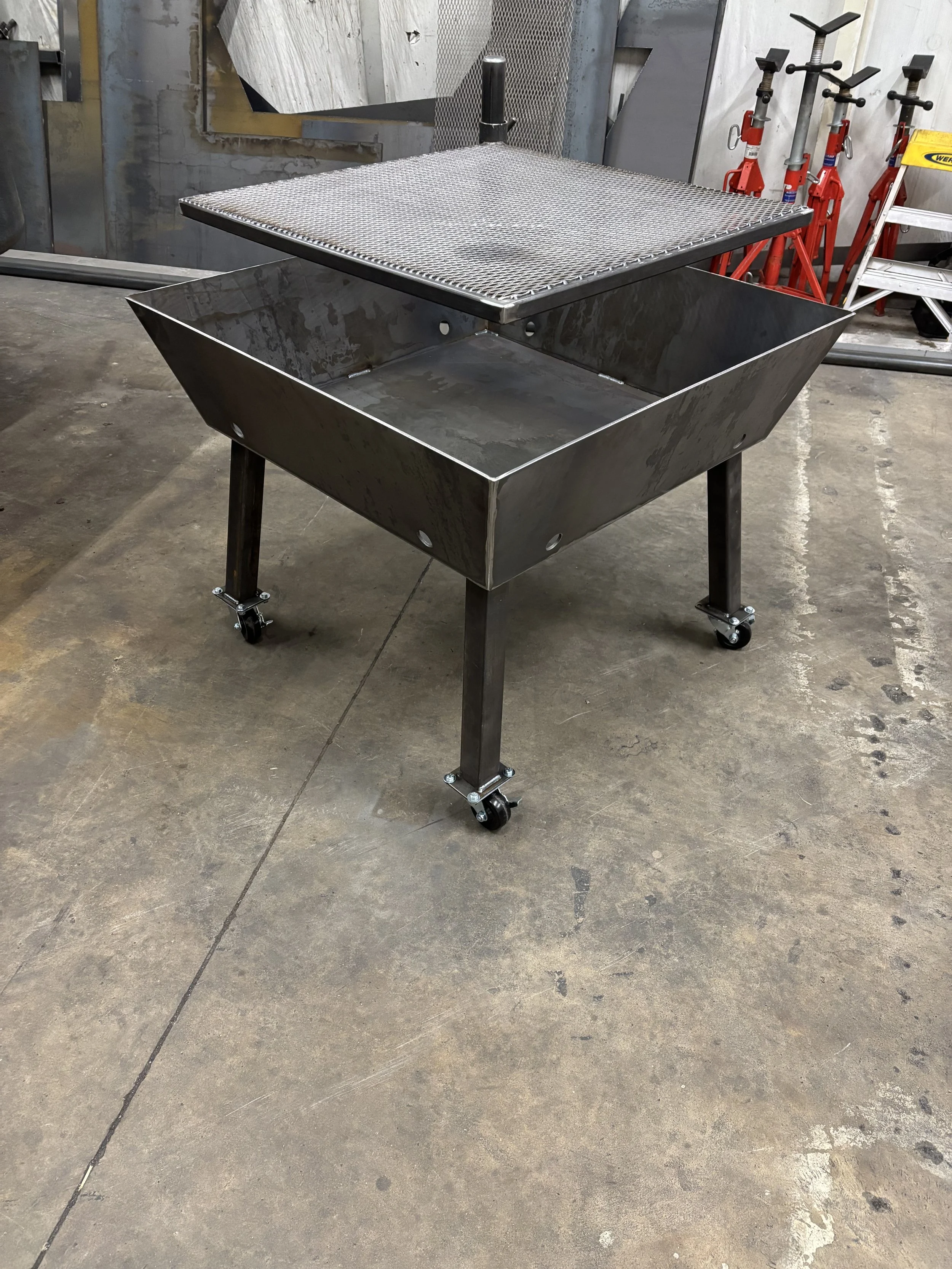 A metal fire pit table by A1 Metal Fabrication, with a mesh lid on wheels, located on a concrete floor in an industrial setting.