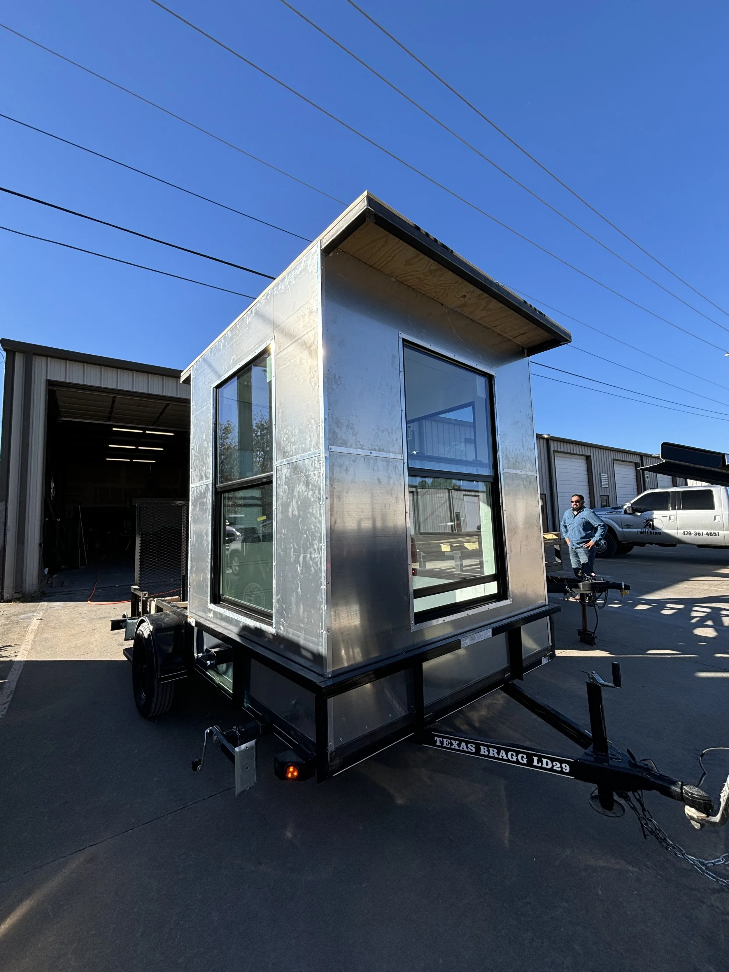 A small metal build by A1 Metal Fabrication with large windows on a trailer, parked outside a warehouse on a clear day.
