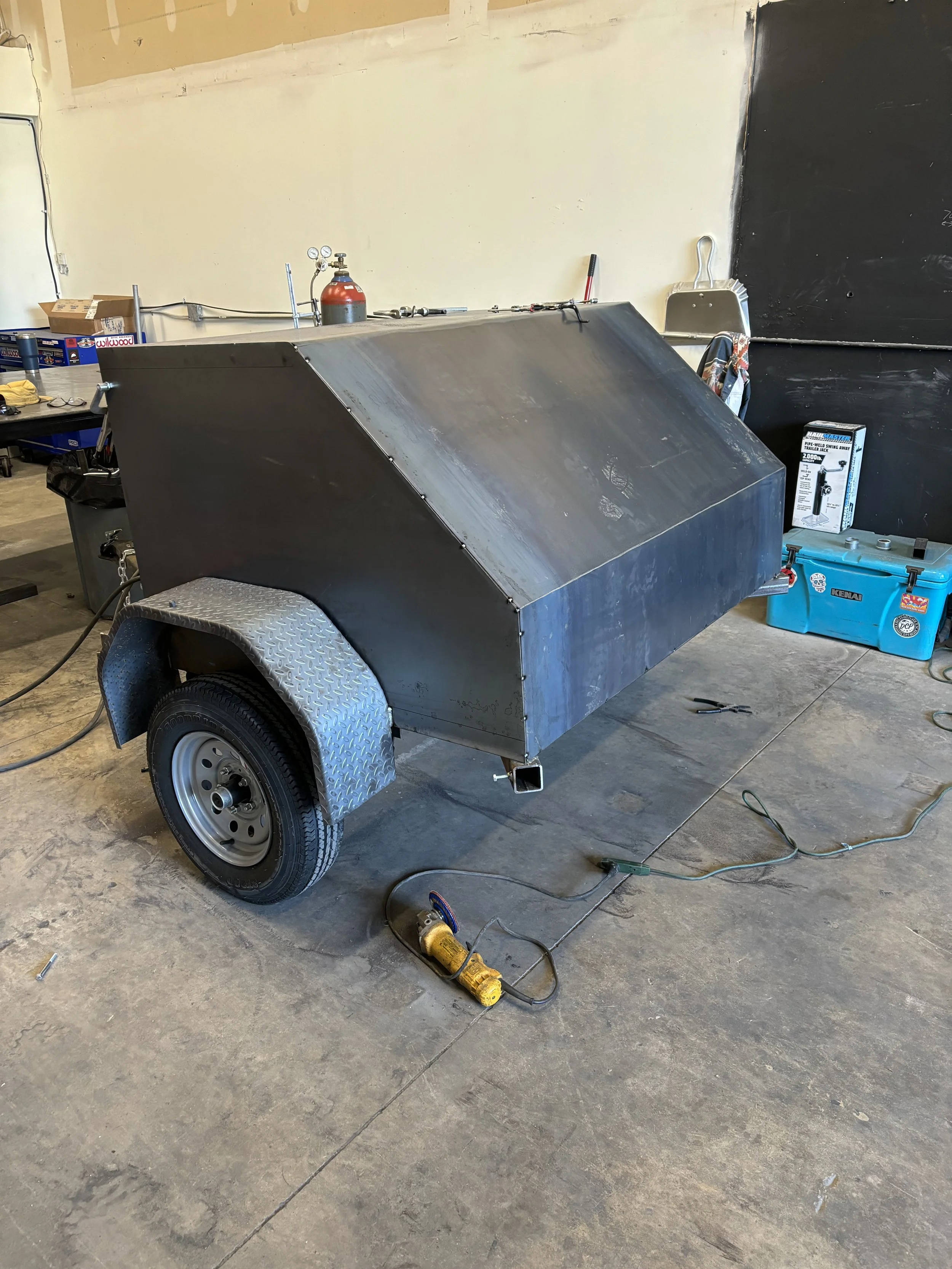 A small welded metal trailer by A1 Metal Fabrication, with a sloped enclosed top, a single tire, and a diamond plate fender in a workshop setting.