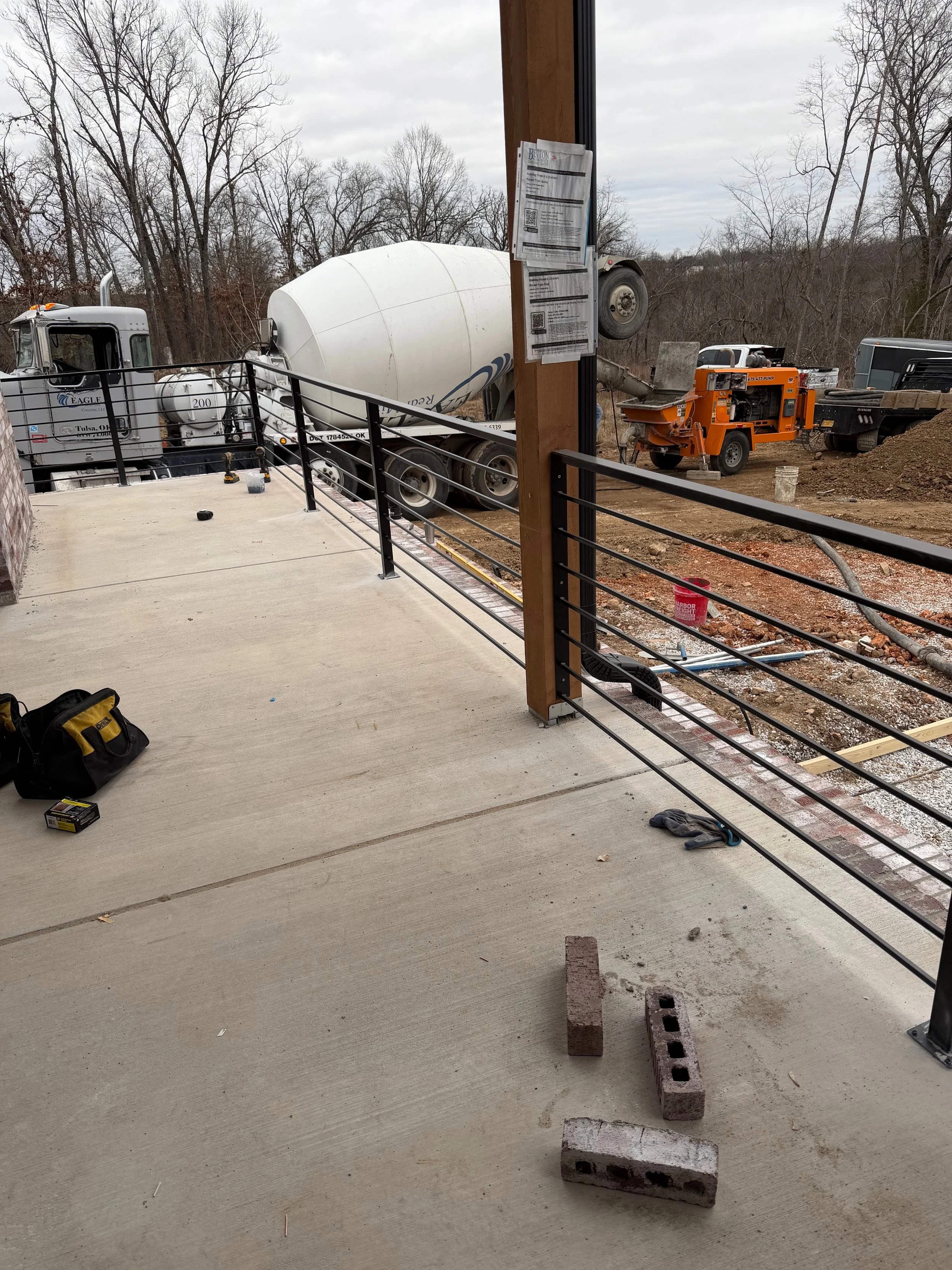 Construction site with a concrete floor, metal railing by A1 Metal Fabrication, and partial building structure. A cement mixer truck is parked outside, and construction equipment and materials are visible around the area.