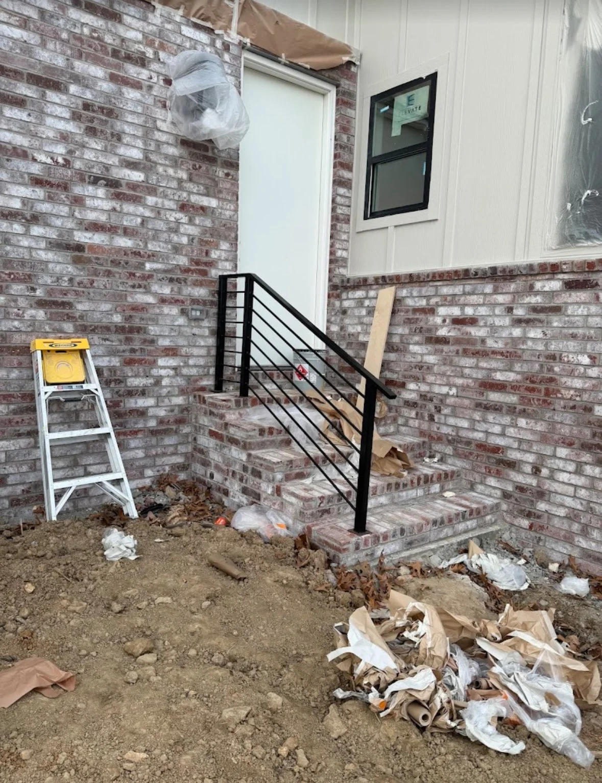 Construction site showing brick steps, black metal railing by A1 Metal Fabrication, interior doorway, and construction debris including paper and plastic on the ground.