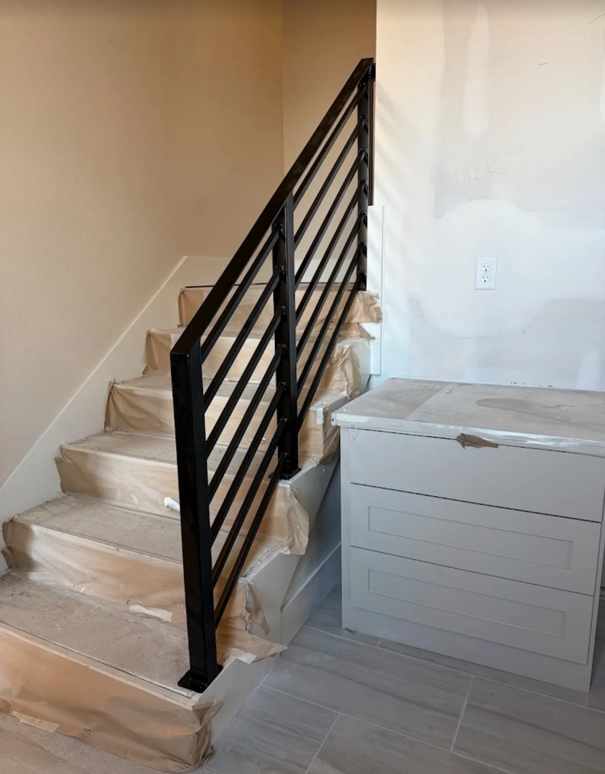 Interior of a house under renovation showing stairs with black metal railing  by A1 Metal Fabrication, partially covered with brown paper and painter's tape, adjacent to a white wall and a cabinet.