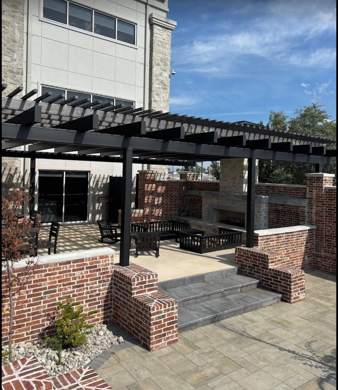 Outdoor patio area with brick walls, a stone fireplace, black outdoor furniture, and a pergola with slatted roof, under a blue sky with some clouds. A1 Metal Fabrication.