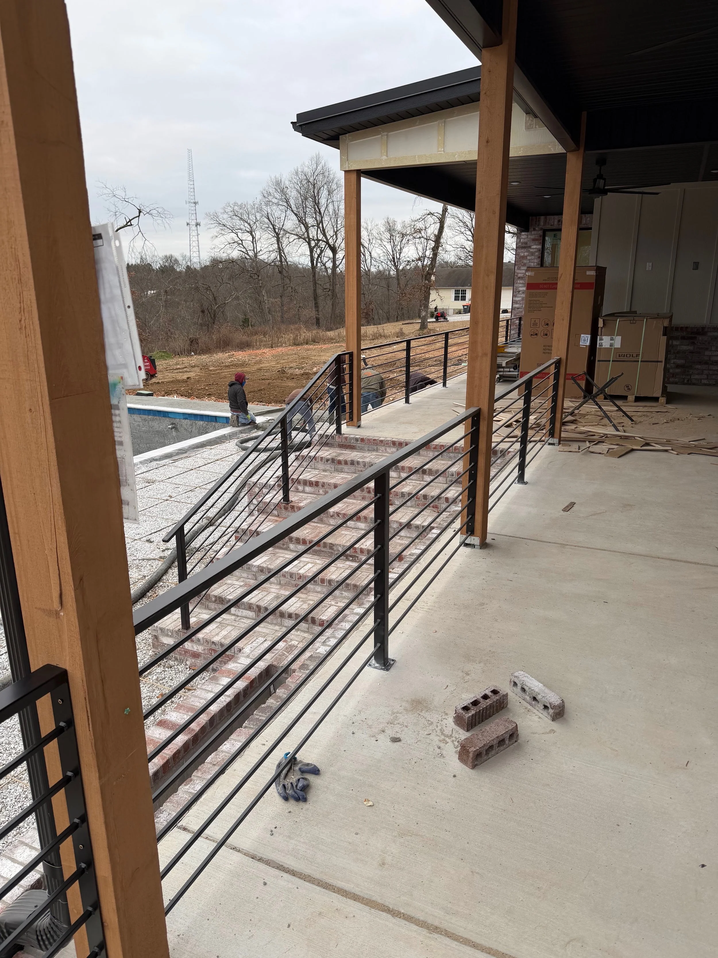Construction site showing a concrete porch with black metal railings by A1 Metal Fabrication, brick stairs, construction tools and bricks on the floor, people working outside, and a wooded area in the background.