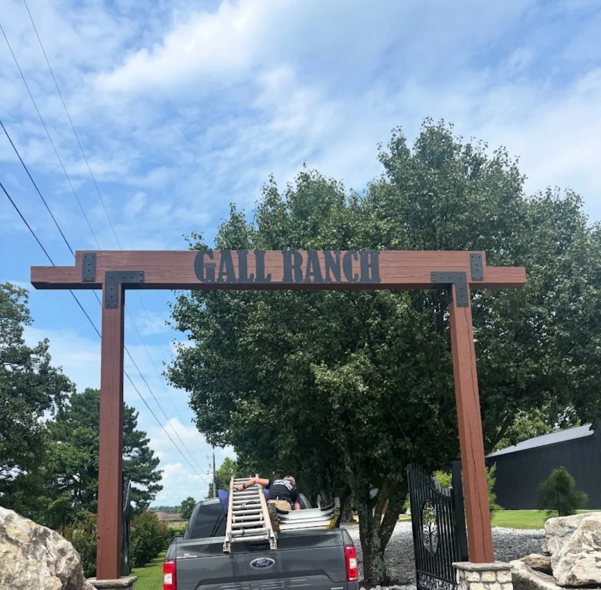 Entrance sign by A1 Metal Fabrication reading 'Gall Ranch' supported by wooden posts, with a truck parked below and workers setting up ladders, surrounded by trees and rocks.