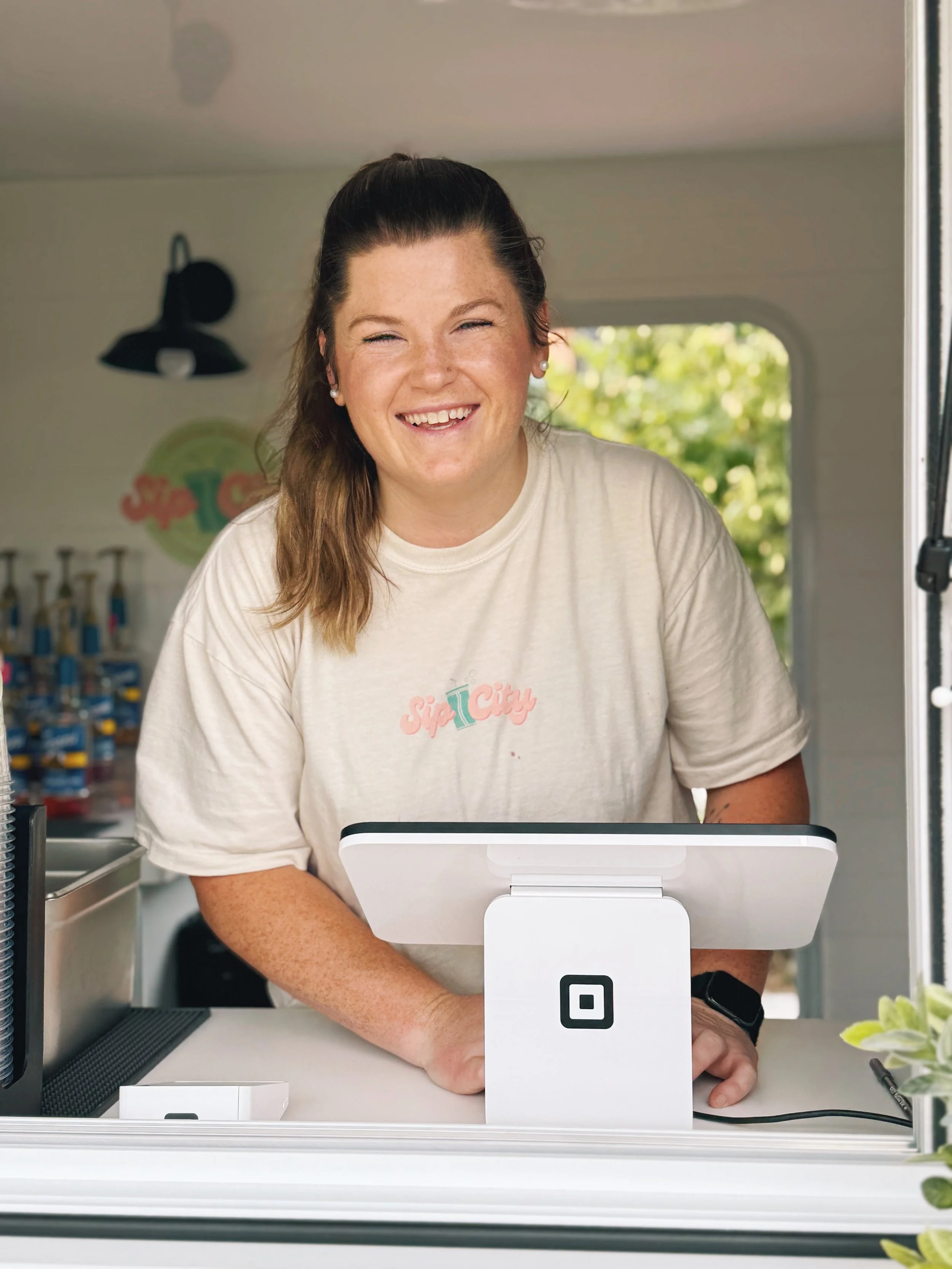 A smiling woman with brown hair in a ponytail, wearing a beige t-shirt with 'Sip City' logo, standing inside a mobile coffee stand with a square Point of Sale device, greenery and snacks in the background.