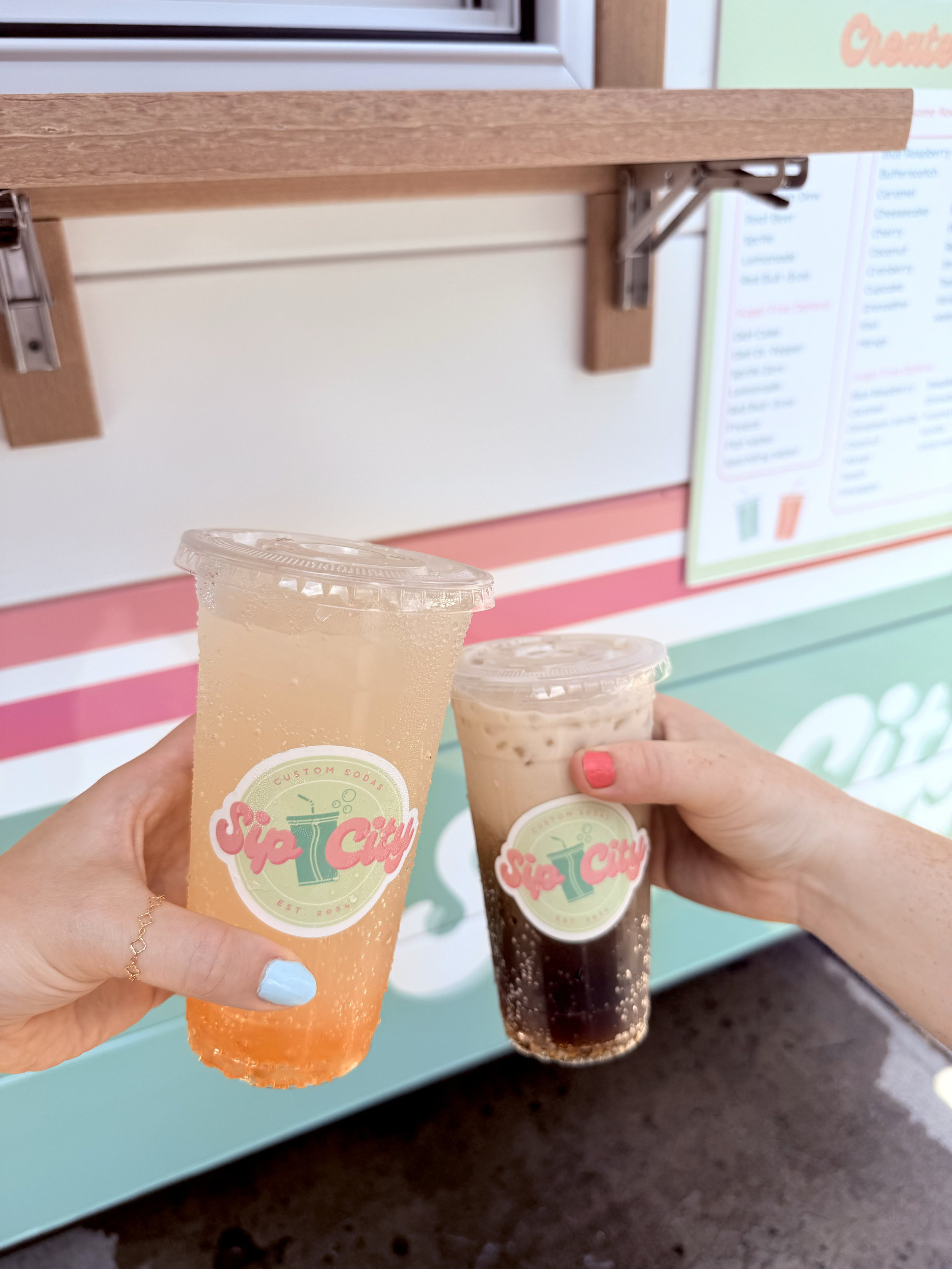 Two hands holding cups of soda, one orange and one dark cola, in front of a sign for Skip City with a colorful background.