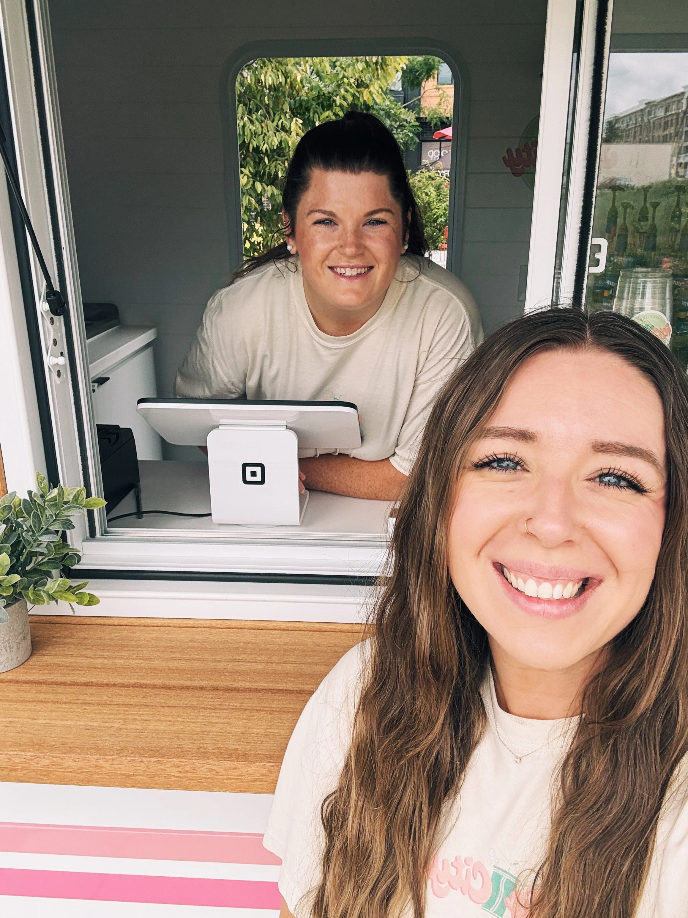 Two women smiling at a selfie inside an ice cream truck, with a counter, a small plant, and a window behind them showing trees and buildings outside.