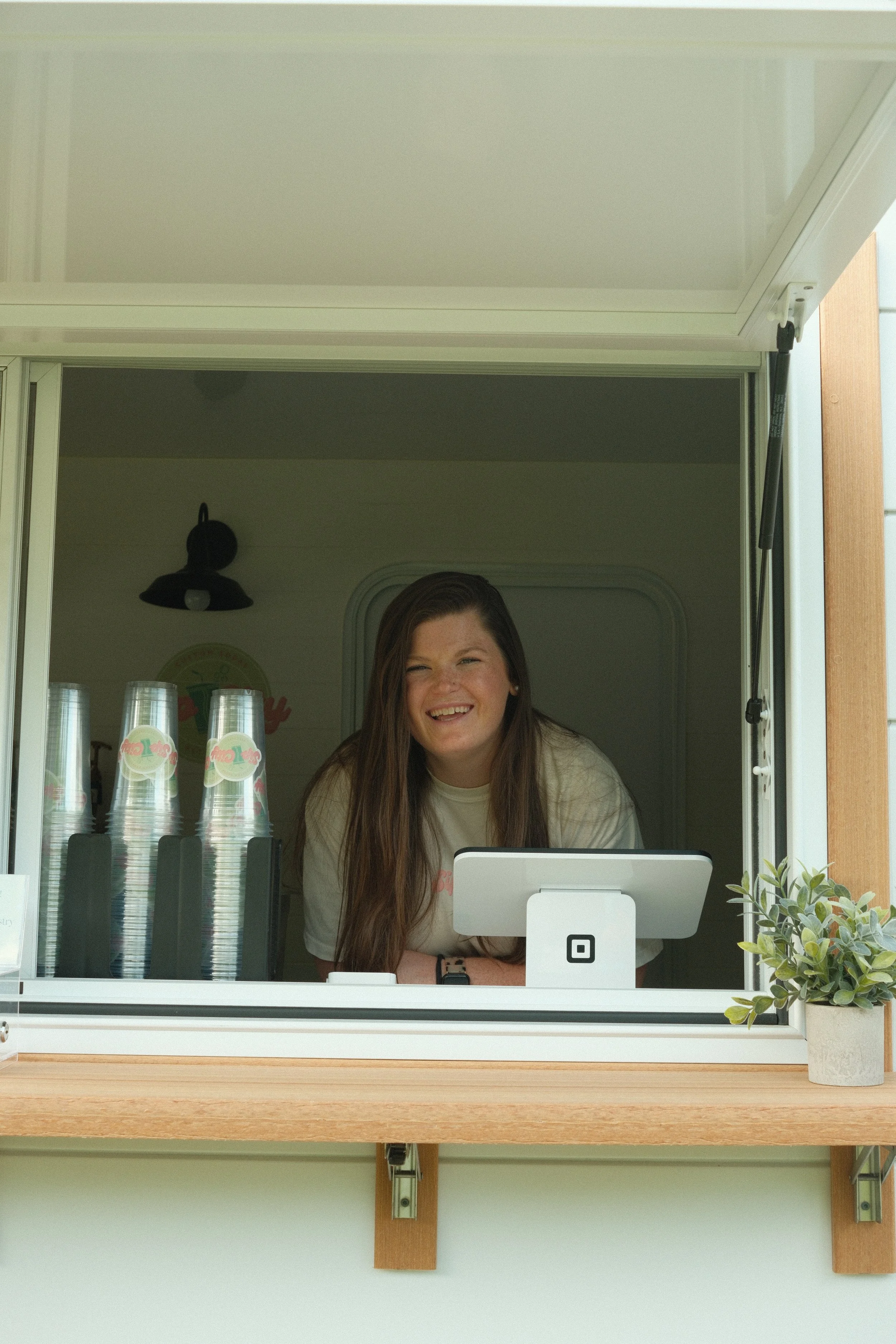 Smiling female employee working at a food truck or kiosk window, with a small plant on the counter.