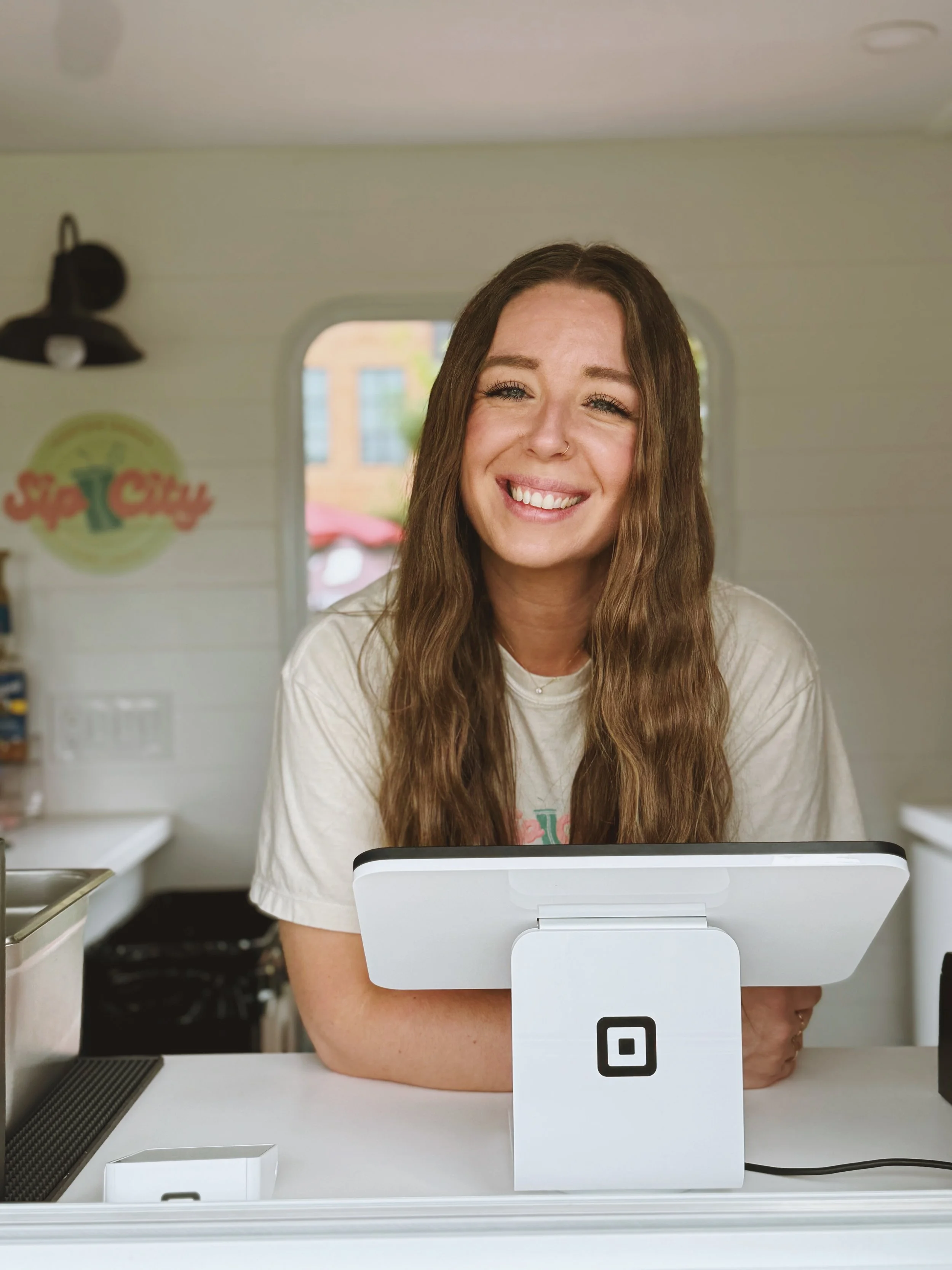 A young woman smiling behind a point-of-sale cash register inside a small shop or food truck.