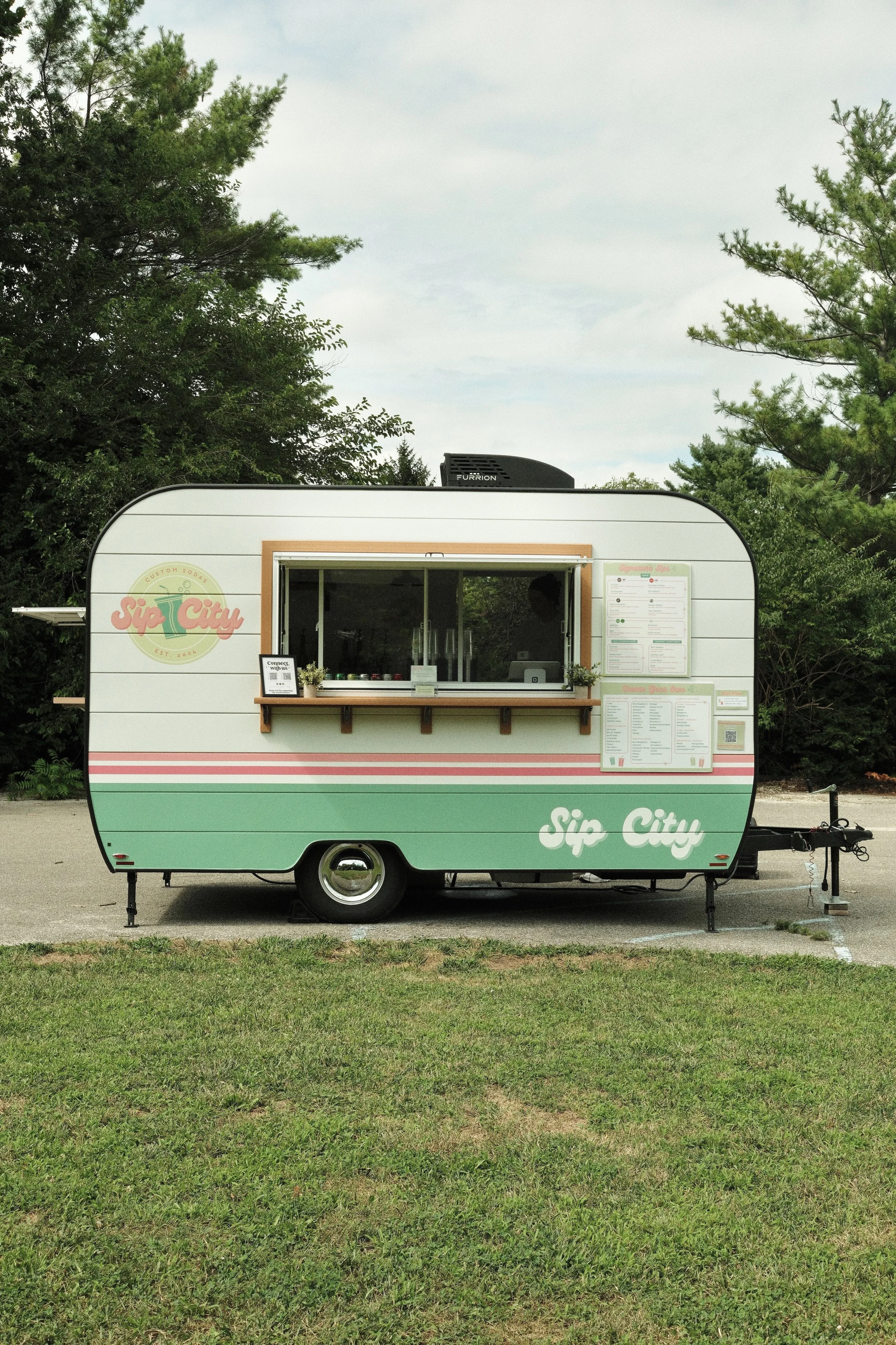 A small mobile food stand called 'Sip City' with a pastel green and pink exterior, serving beverages, parked on a paved lot with trees in the background.