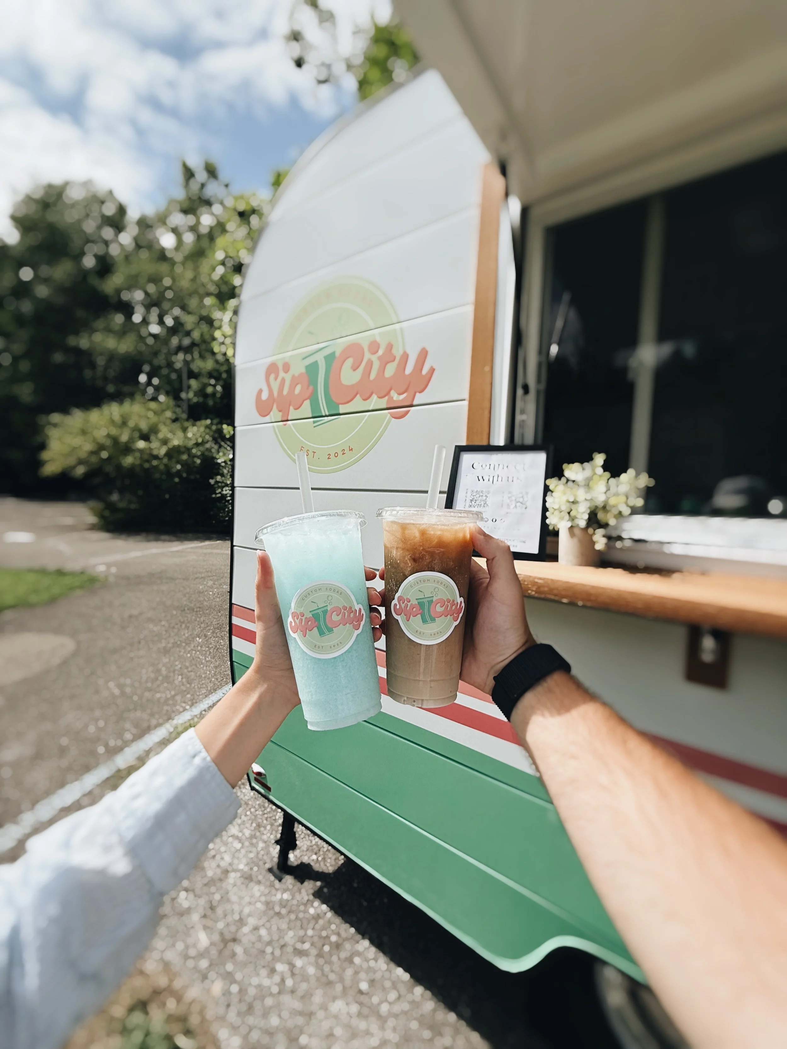 Two people holding iced drinks in front of a vintage-style mobile drink cart with a 'Sip City' logo.