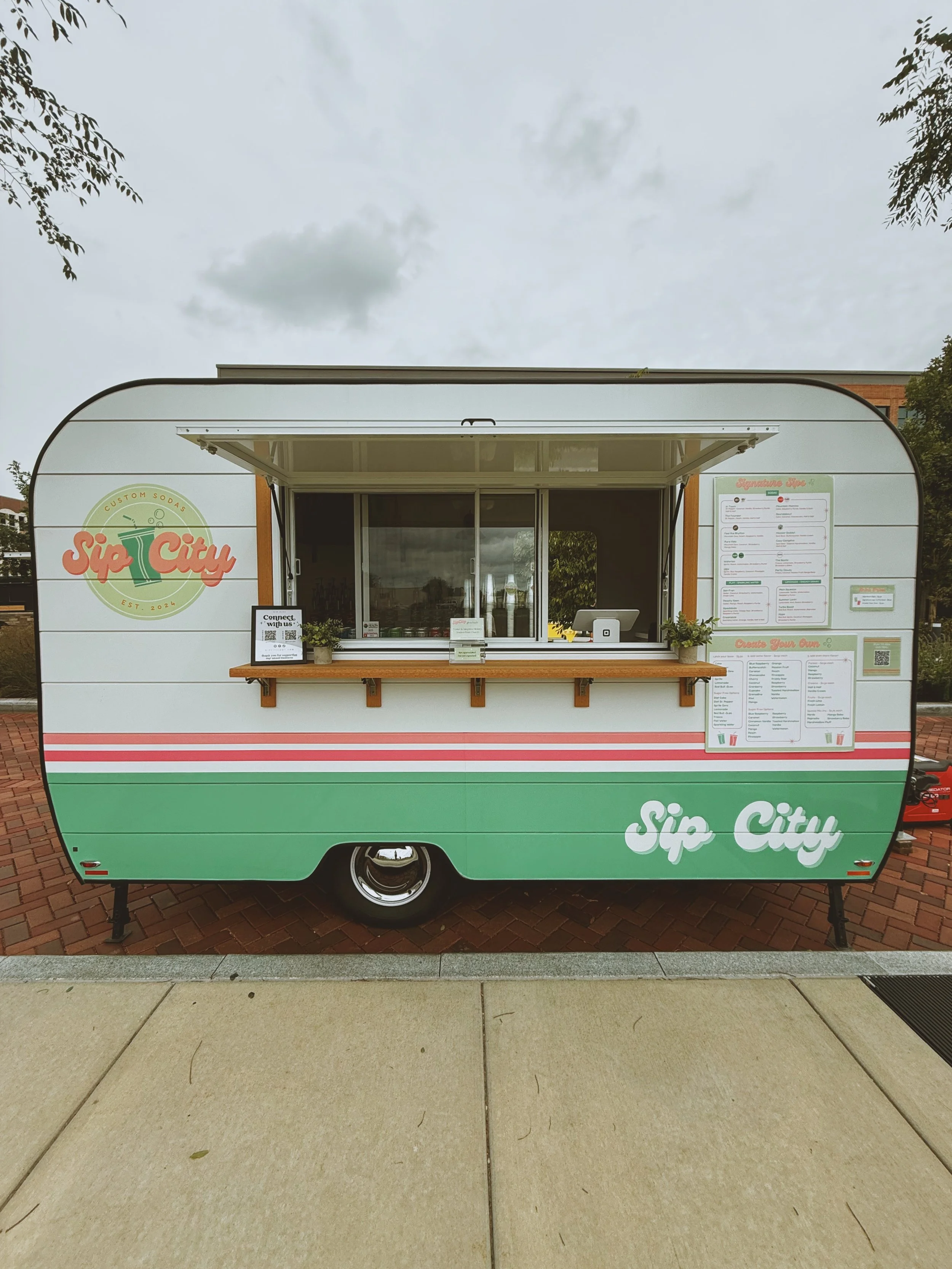 A food truck named Sip City with a pastel green and white exterior, red stripes, and a large menu on the side. The truck has a serving window with a small counter and a cash register.