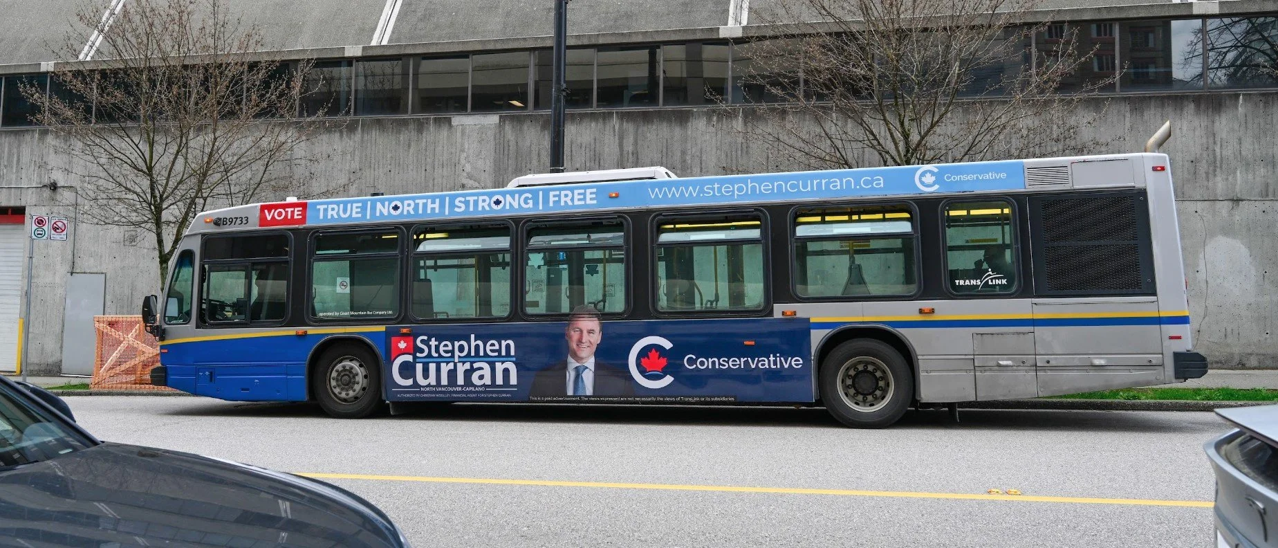 A transit bus with campaign advertisements for Stephen Curran, a Conservative candidate, parked on a city street with houses and trees in the background.