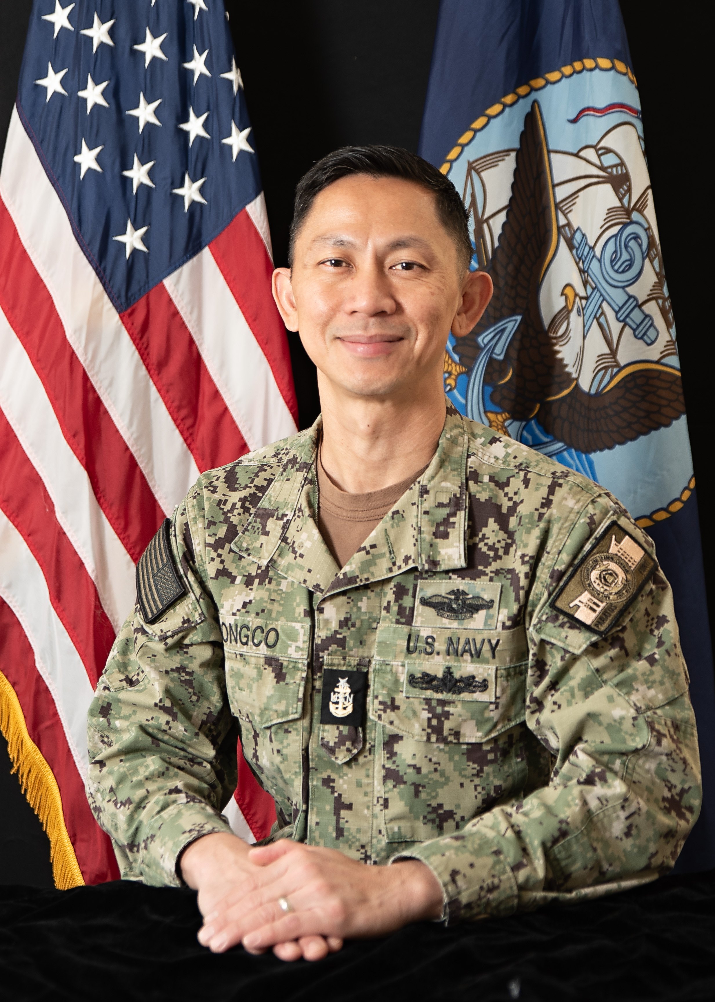 A man in U.S. Navy camouflage uniform sitting in front of American and Navy flags, smiling at the camera.