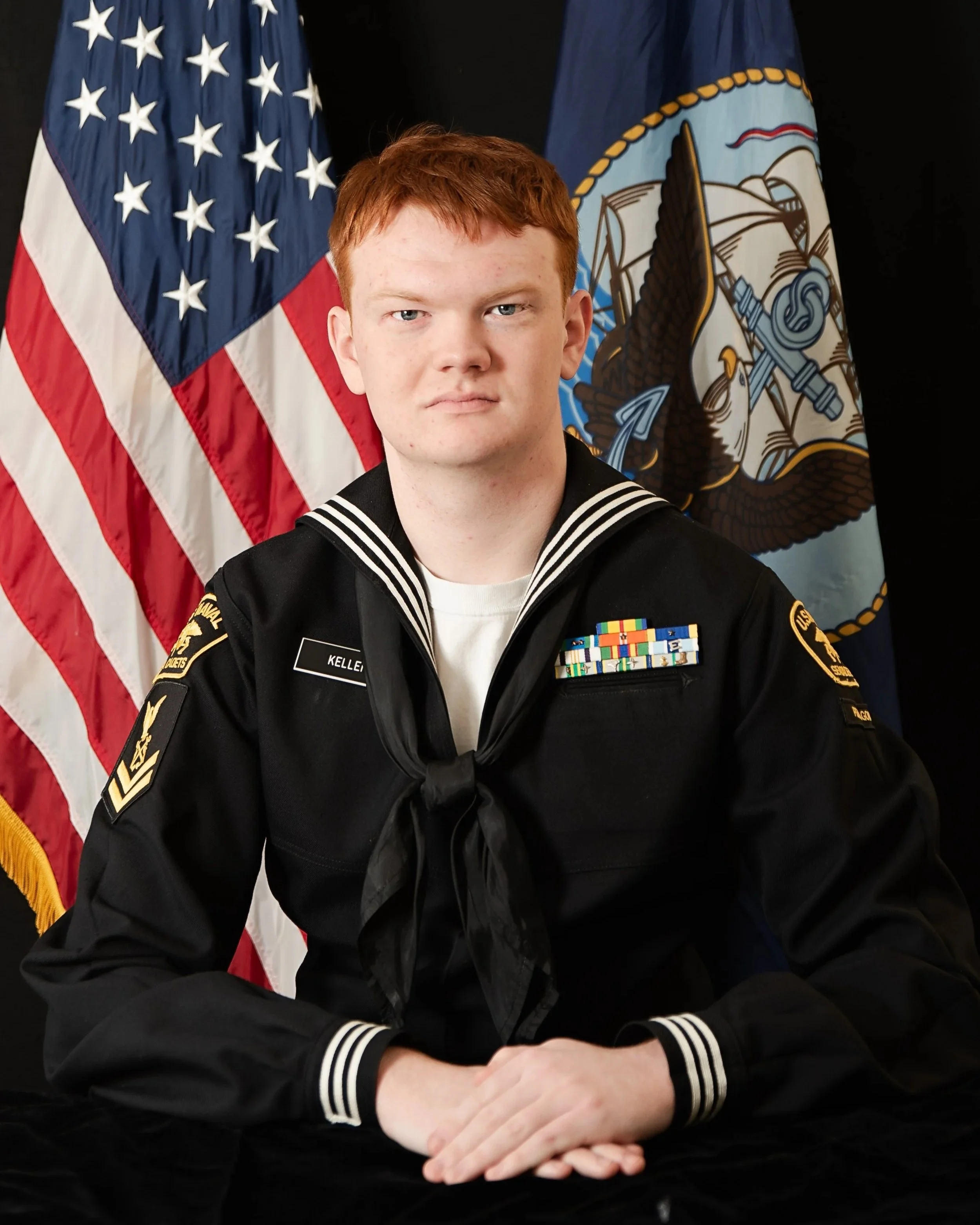 A young man in a USNSCC uniform sitting at a desk with American and military flags behind him.