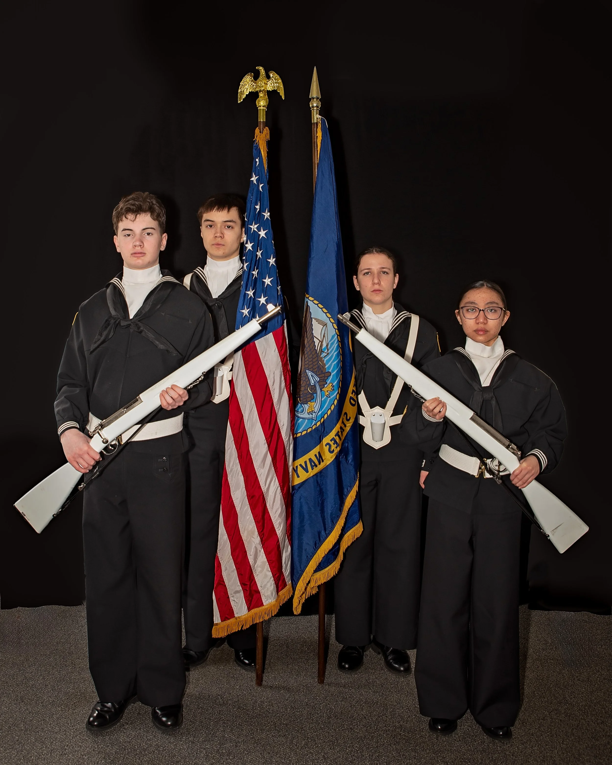 Four young sailors in uniform standing in front of a black background, holding rifles, and flanked by the American flag and the Navy flag.