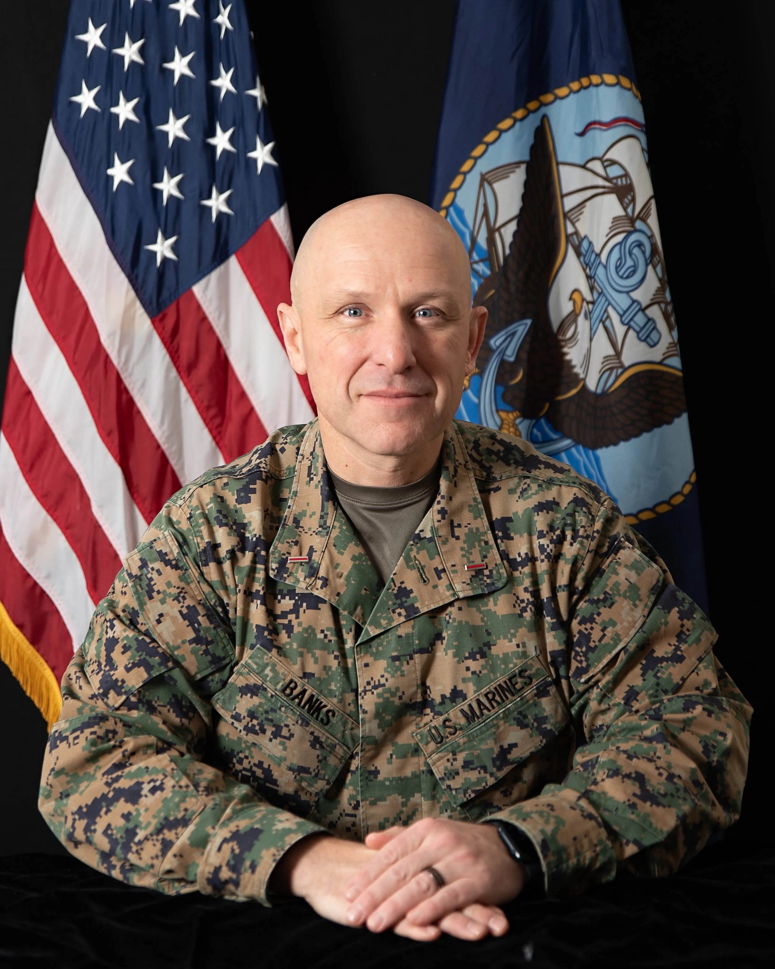 A U.S. Marine in camouflage uniform sitting at a desk, with the American flag and the Navy flag in the background.