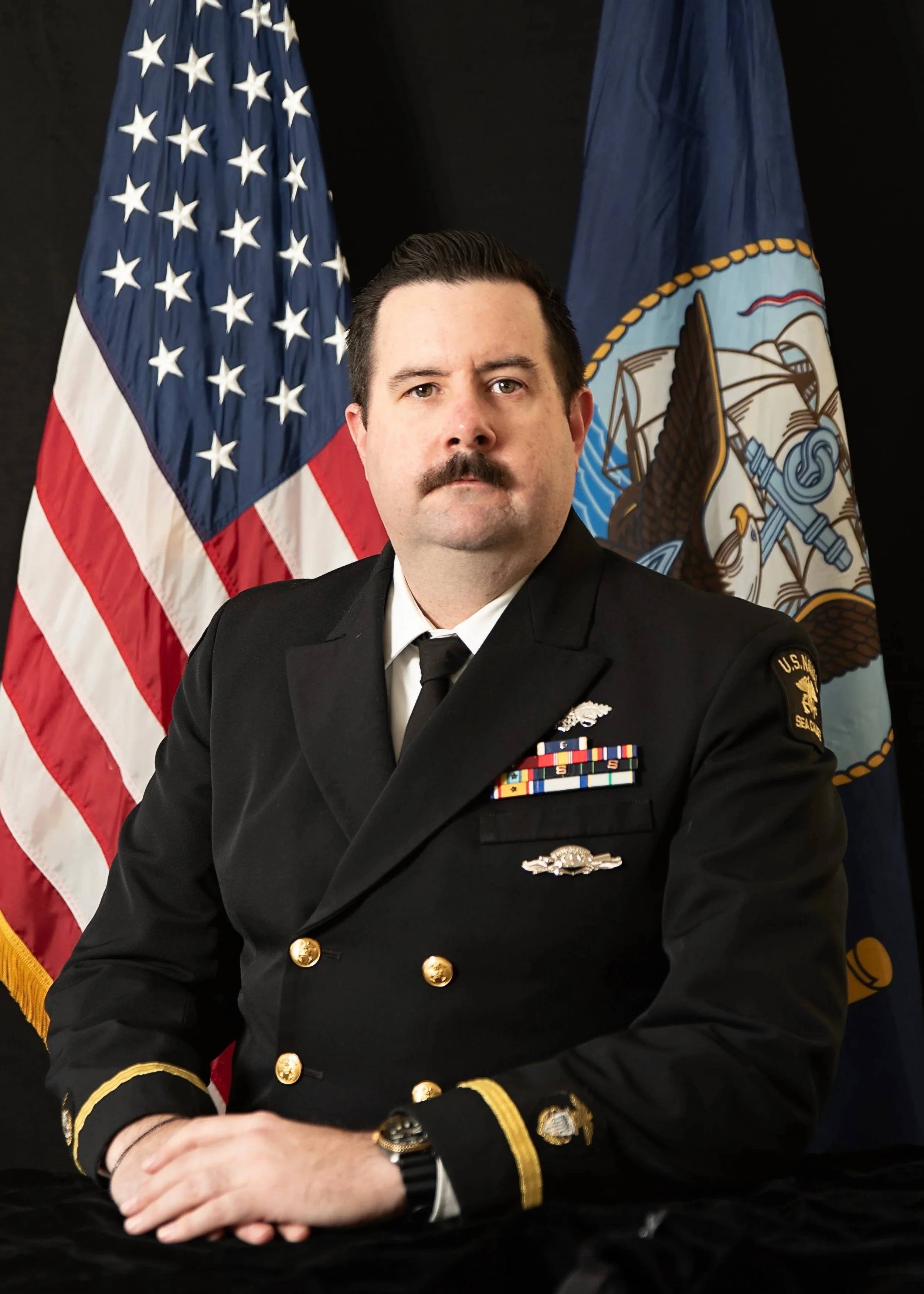 A USNSCC officer in uniform sitting with hands clasped in front of him, American flag and navy flag in the background.