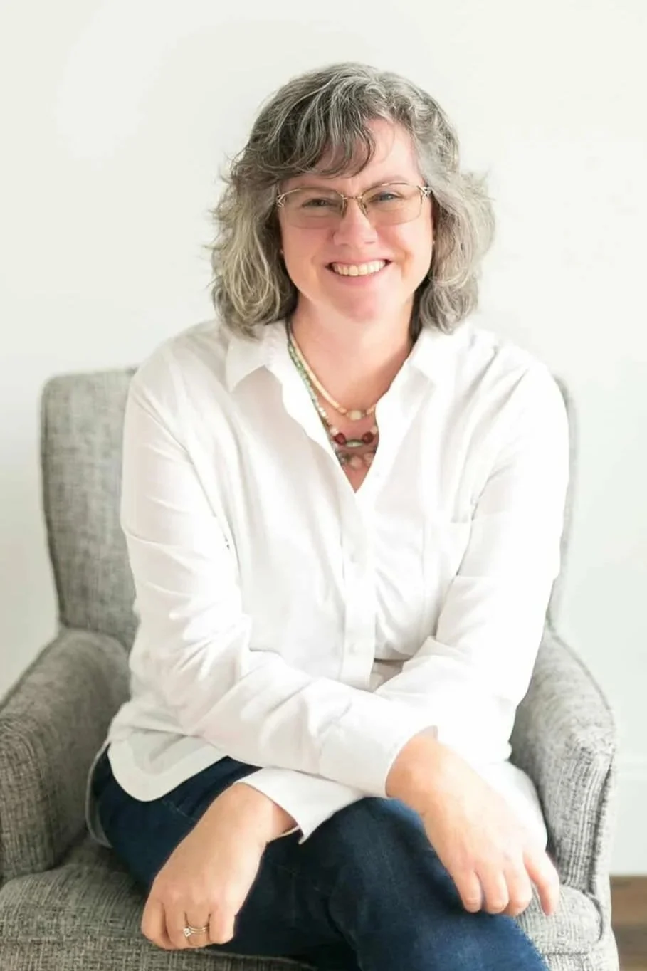 A woman with gray curly hair and glasses smiling, sitting on a gray armchair against a plain white background, wearing a white shirt and dark jeans.