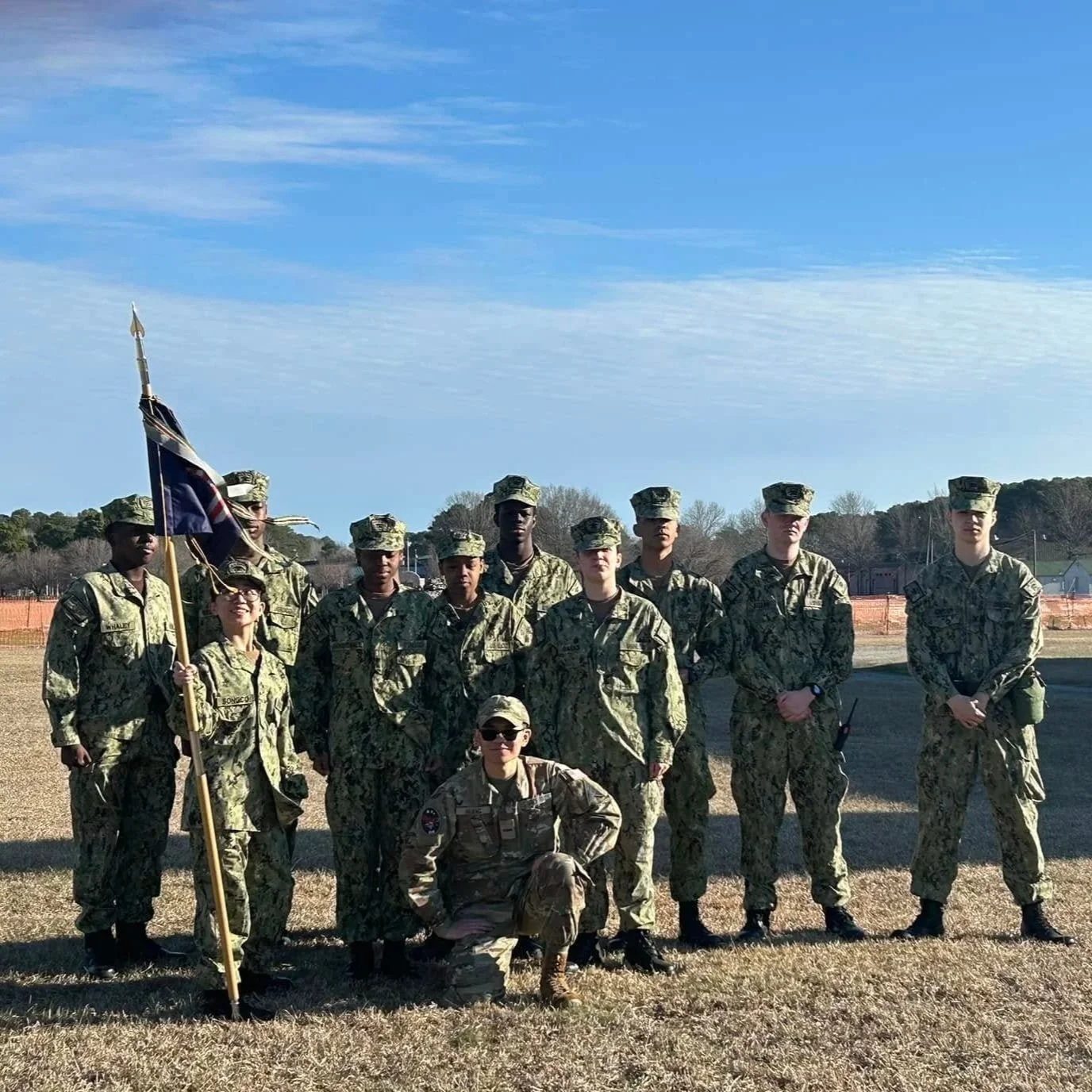 A group of soldiers in camouflage uniforms posing outdoors, some standing and one kneeling in front, holding a flag.