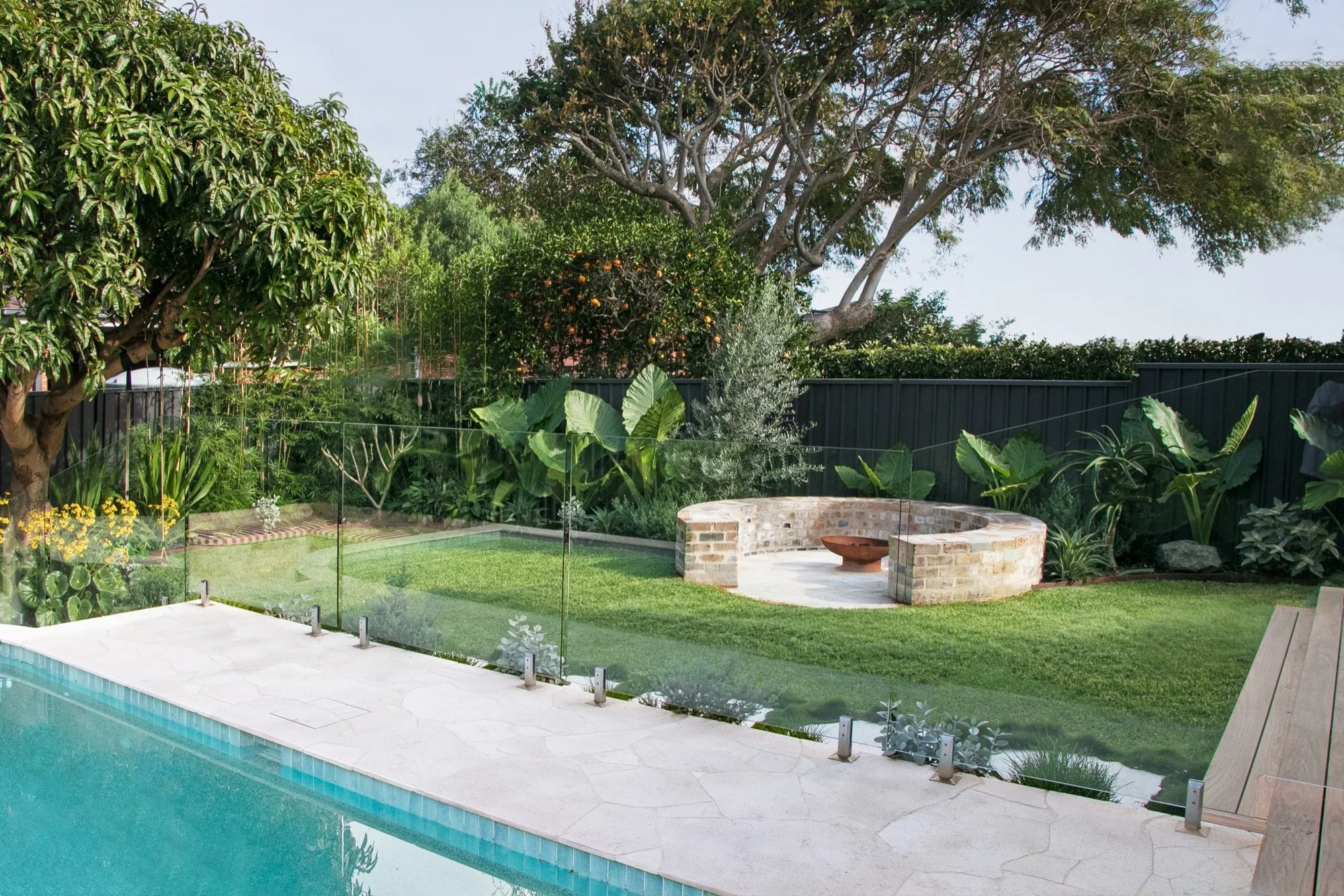Pool coping and glass fence looking across the lawn to a recycled brick fire pit in Chifley