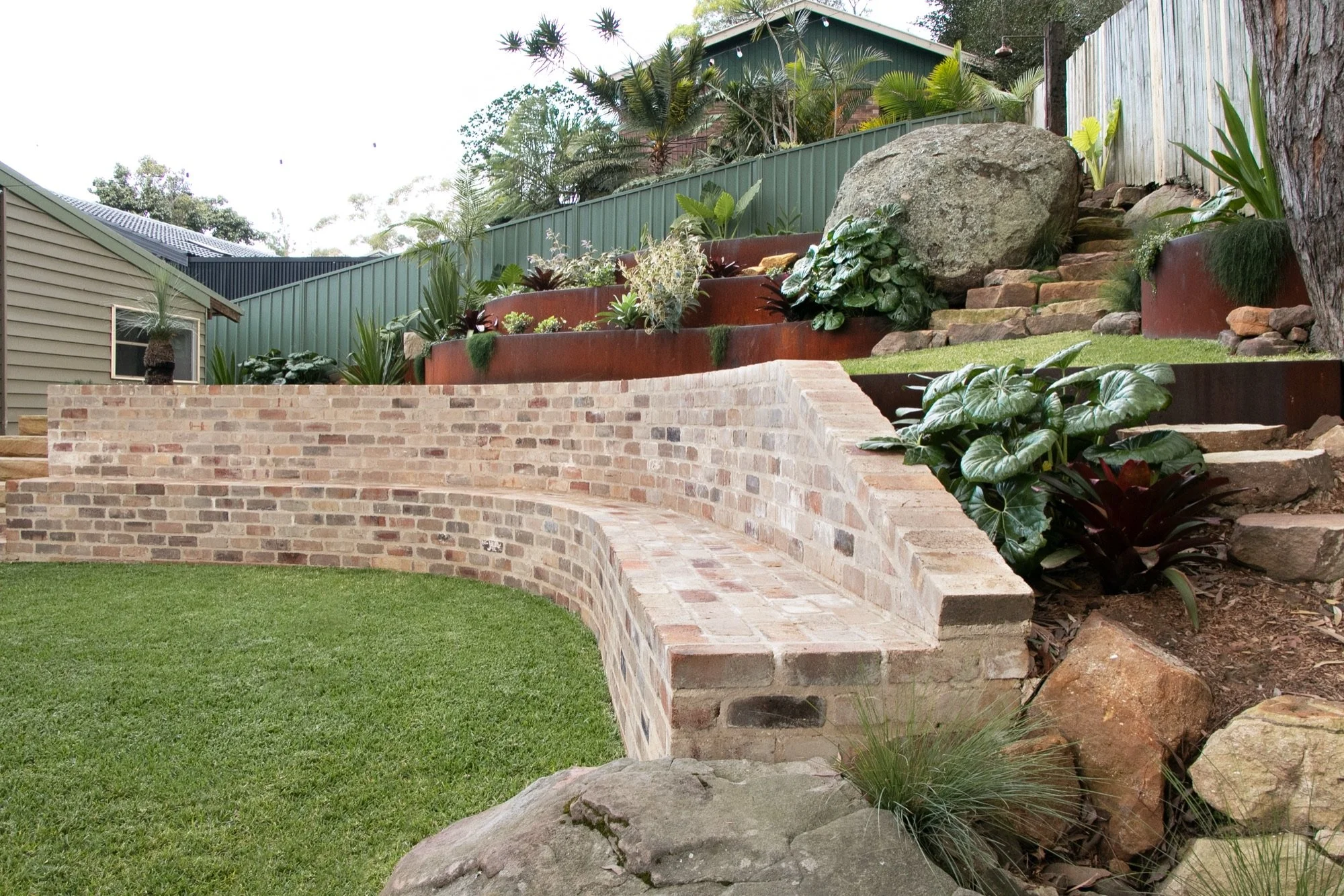 Curved recycled brick bench and retaining wall wrapping a level lawn in a terraced Bulli garden