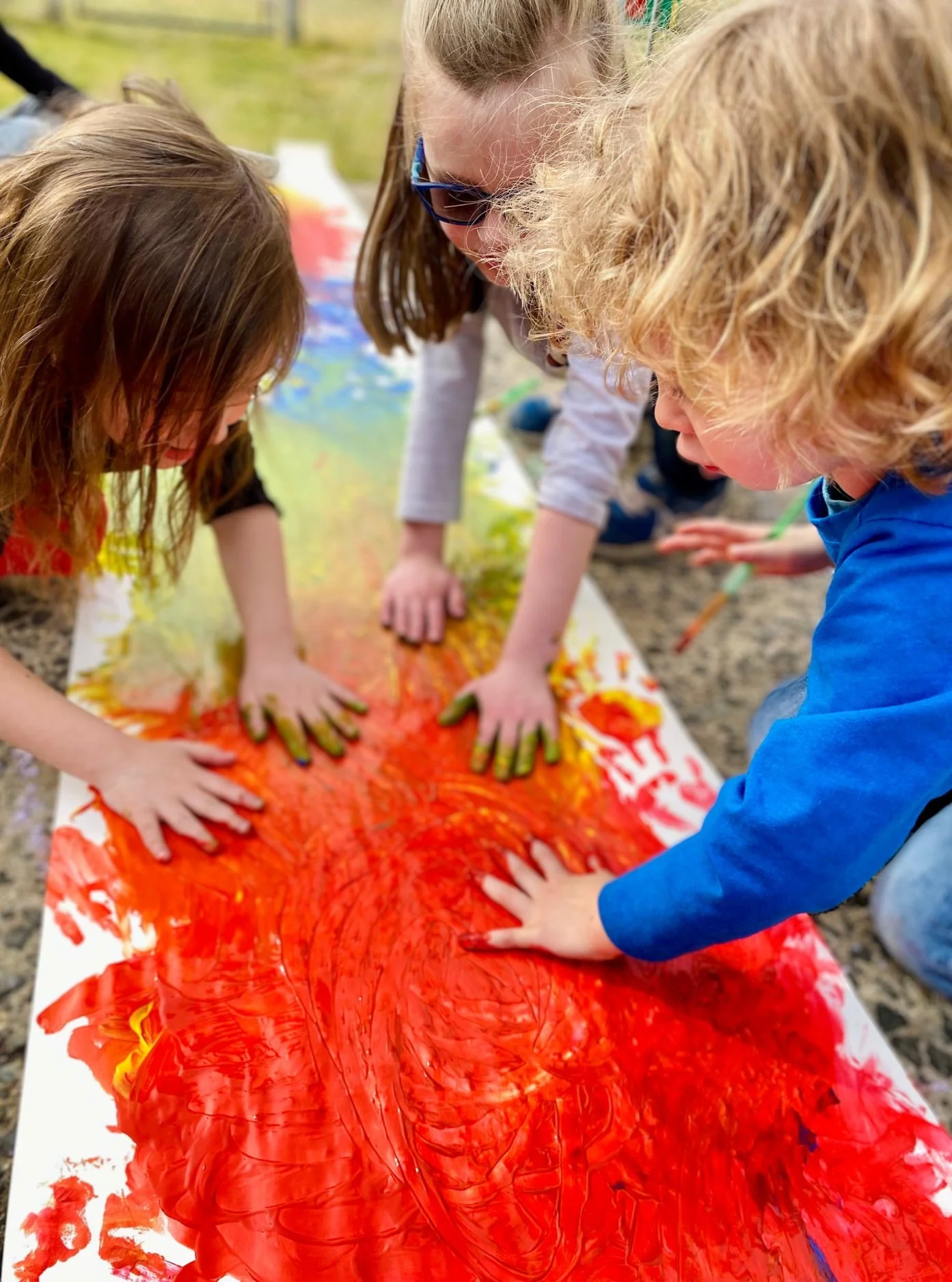 Children with paint-covered hands engaging in finger painting outdoors on a long sheet of paper.
