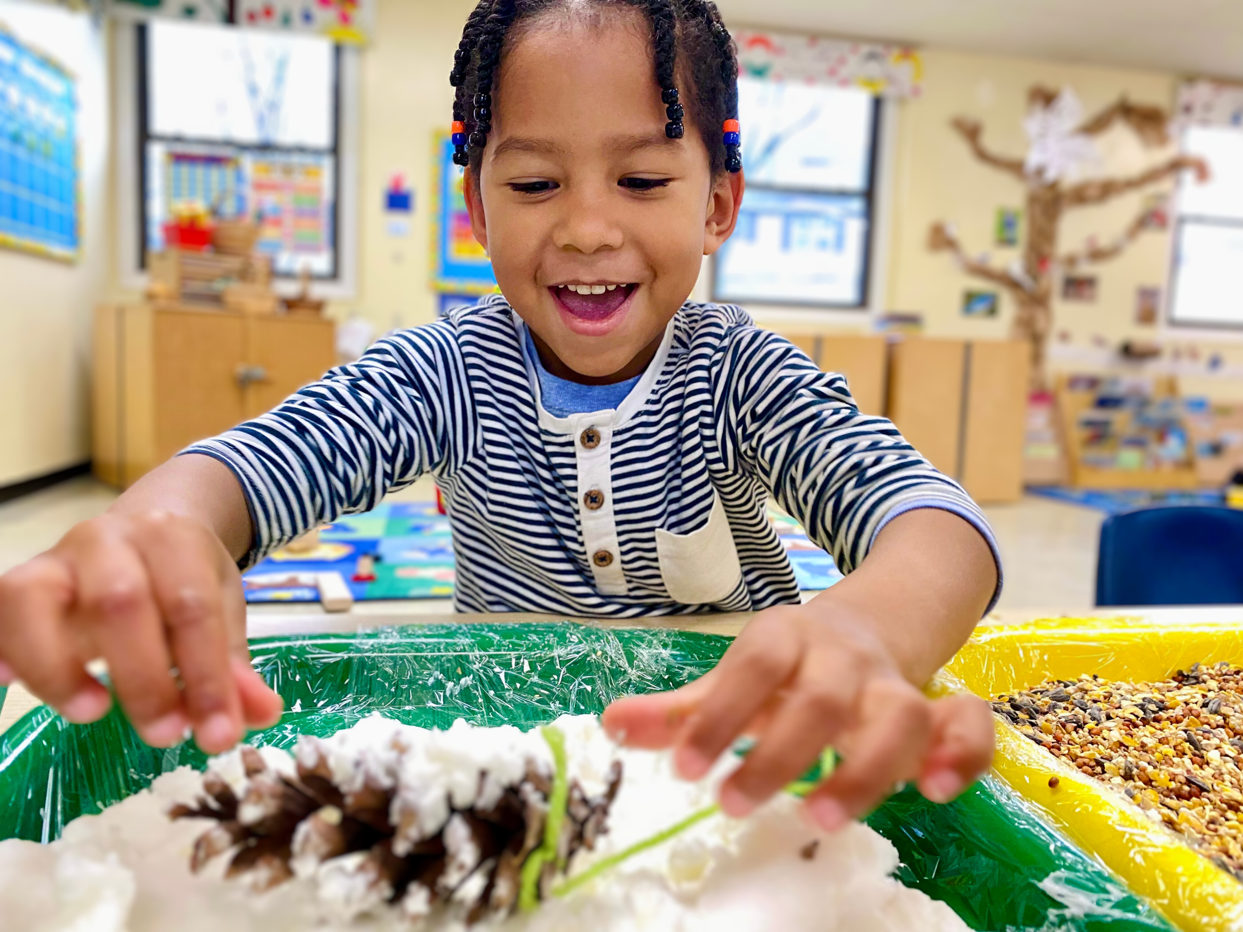 Young boy in a striped shirt playing with a sensory or craft activity at a classroom table, smiling and focused.